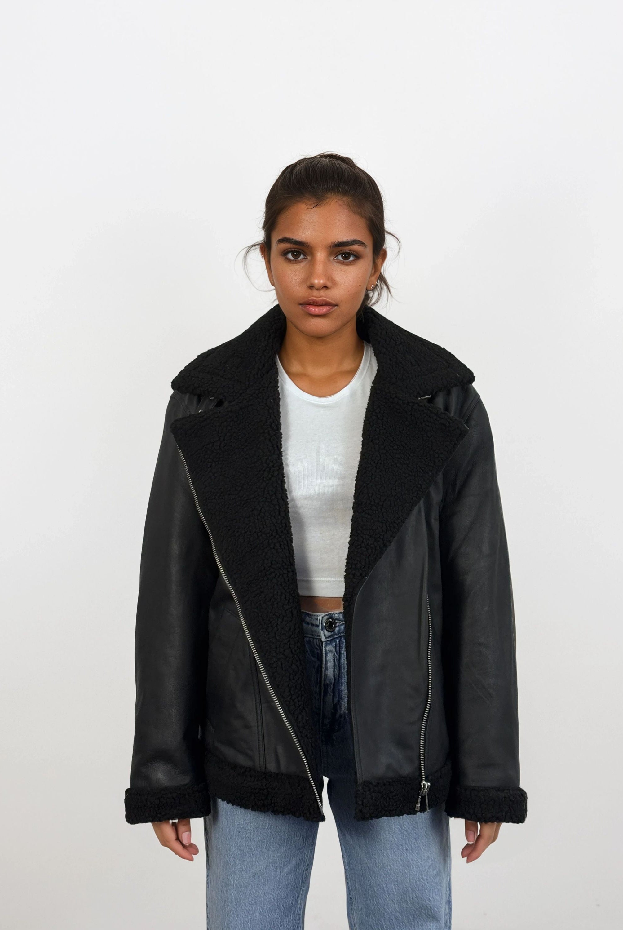 Eine Frau trägt vor weißem Hintergrund eine schwarze trueprodigy Damen Lederjacke aus Schafsleder mit Teddy-Besatz über einem weißen Crop-Top und hellblauen Jeans, steht entspannt mit neutralem Gesichtsausdruck.
