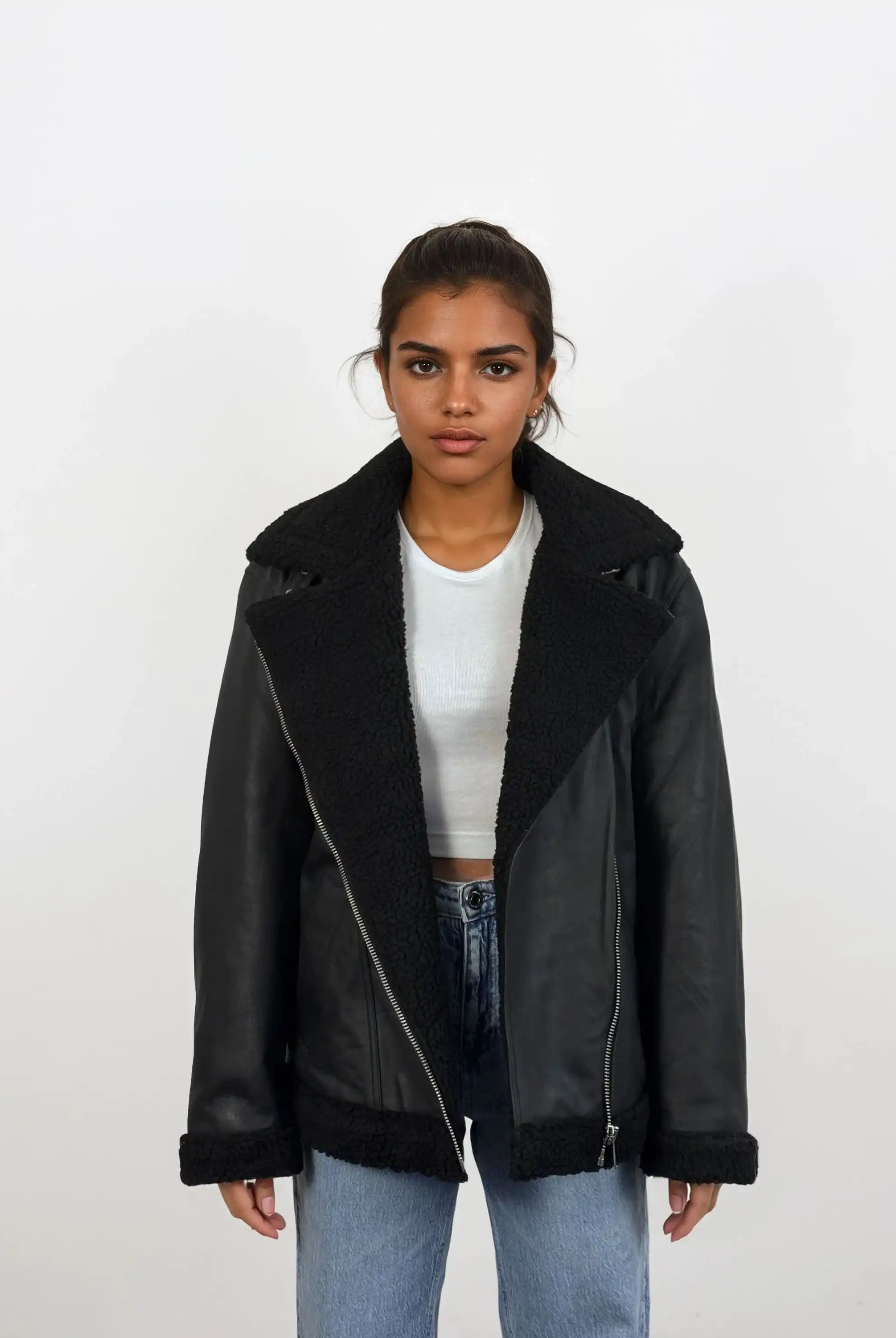 Eine Frau trägt vor weißem Hintergrund eine schwarze trueprodigy Damen Lederjacke aus Schafsleder mit Teddy-Besatz über einem weißen Crop-Top und hellblauen Jeans, steht entspannt mit neutralem Gesichtsausdruck.