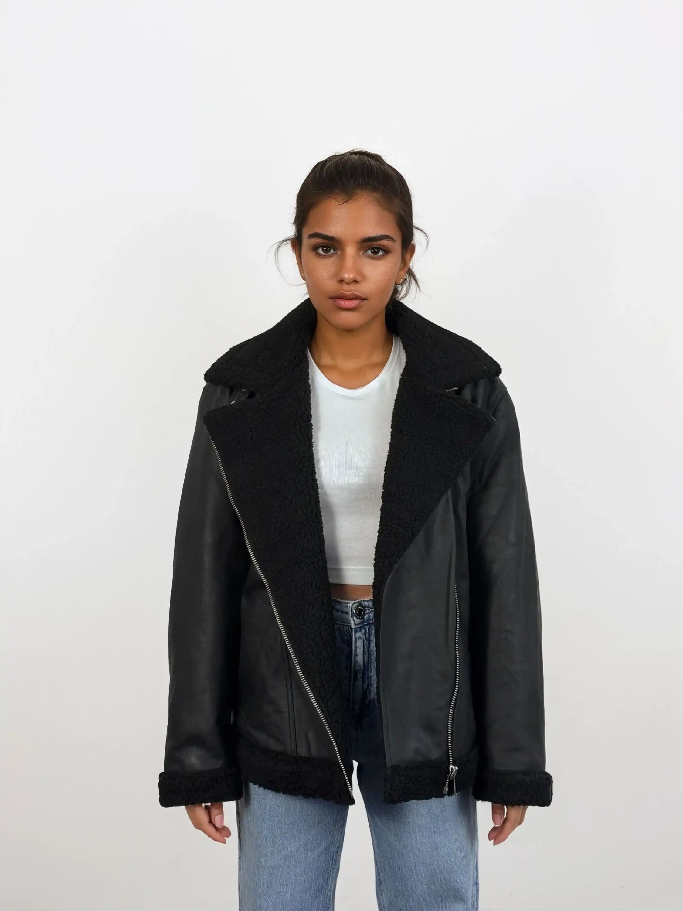 Eine Frau trägt vor weißem Hintergrund eine schwarze trueprodigy Damen Lederjacke aus Schafsleder mit Teddy-Besatz über einem weißen Crop-Top und hellblauen Jeans, steht entspannt mit neutralem Gesichtsausdruck.