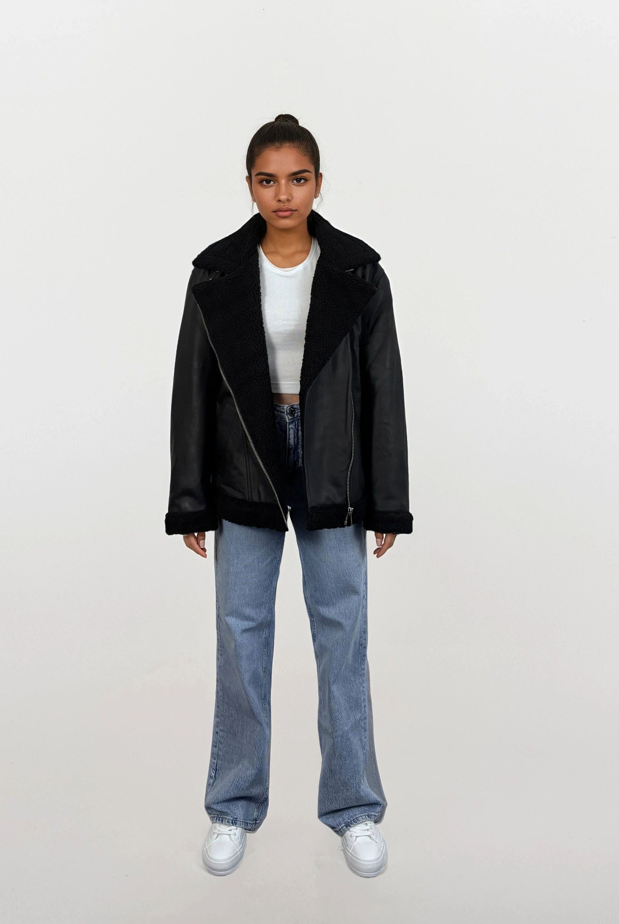 Eine Person steht frontal und trägt die trueprodigy Damen Lederjacke aus Schafsleder in Schwarz mit Teddy-Besatz, ein weißes Cropped Top, locker geschnittene blaue Jeans und weiße Sneaker vor schlicht hellem Hintergrund.
