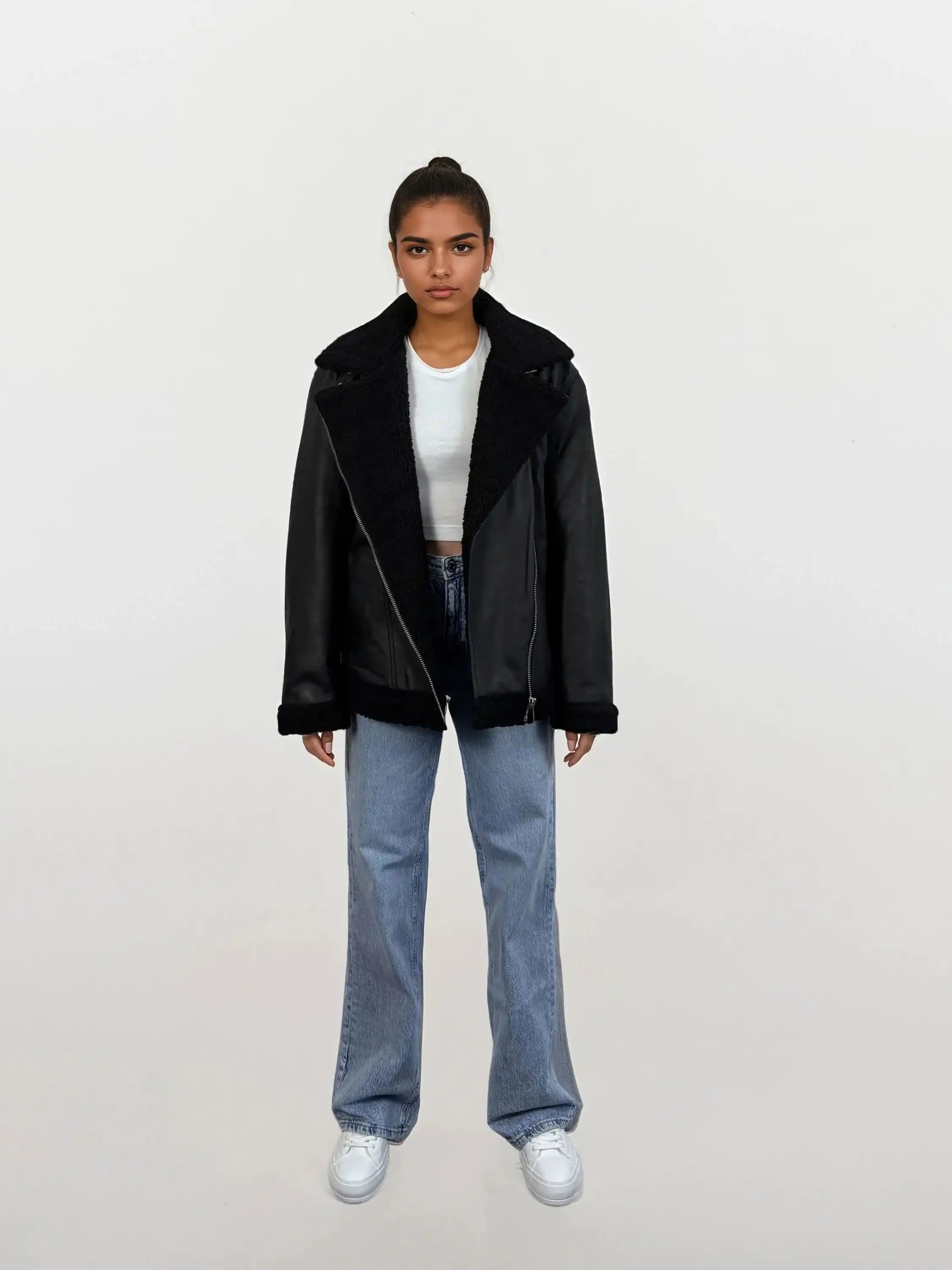 Eine Person steht frontal und trägt die trueprodigy Damen Lederjacke aus Schafsleder in Schwarz mit Teddy-Besatz, ein weißes Cropped Top, locker geschnittene blaue Jeans und weiße Sneaker vor schlicht hellem Hintergrund.