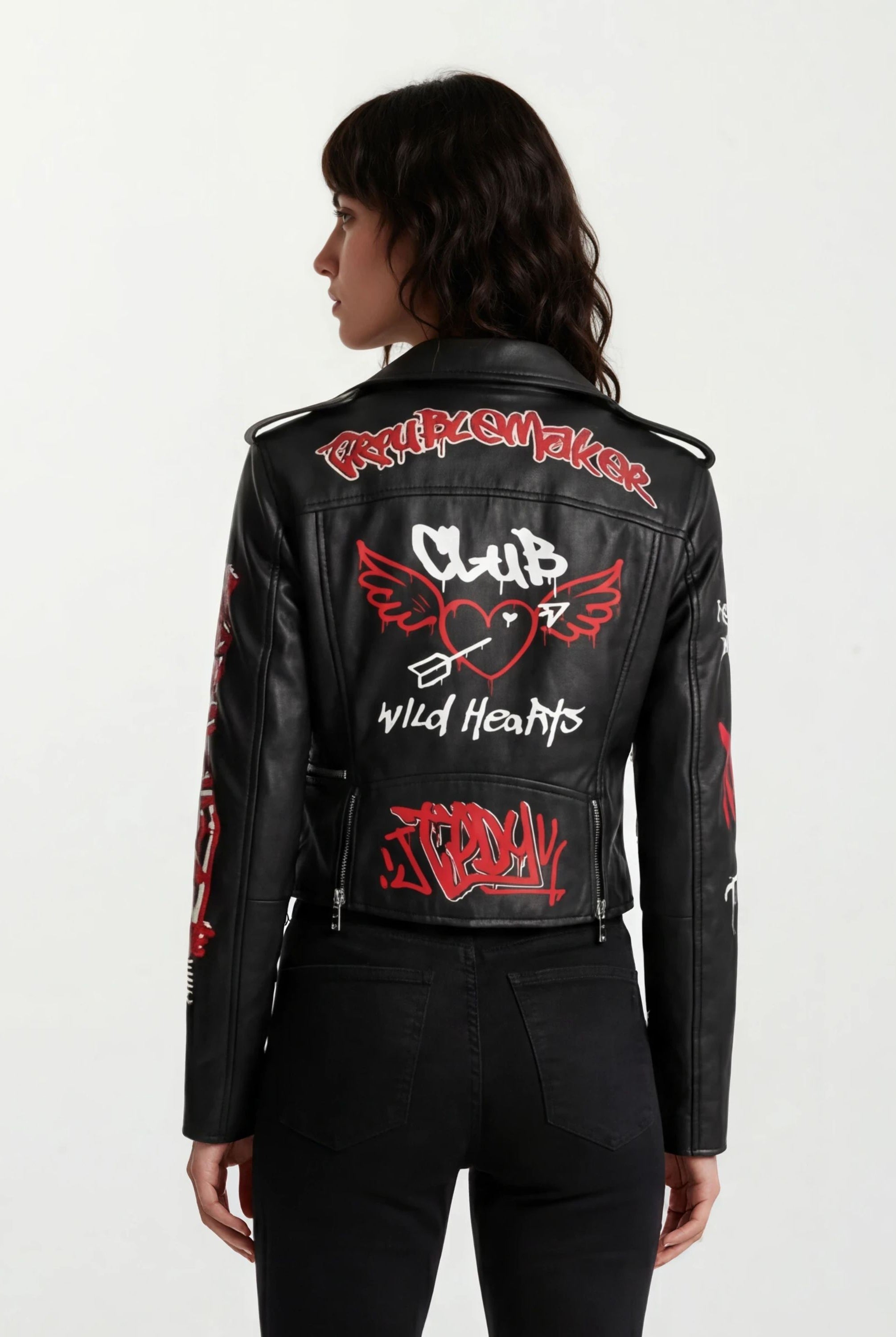 Eine Frau mit mittellangen dunklen Haaren ist von hinten zu sehen. Sie trägt die trueprodigy Feminine Lederjacke mit Grafittiprint und Reißverschluss, eine schwarze Lederjacke mit roten und weißen Graffiti-Grafiken und Text.
