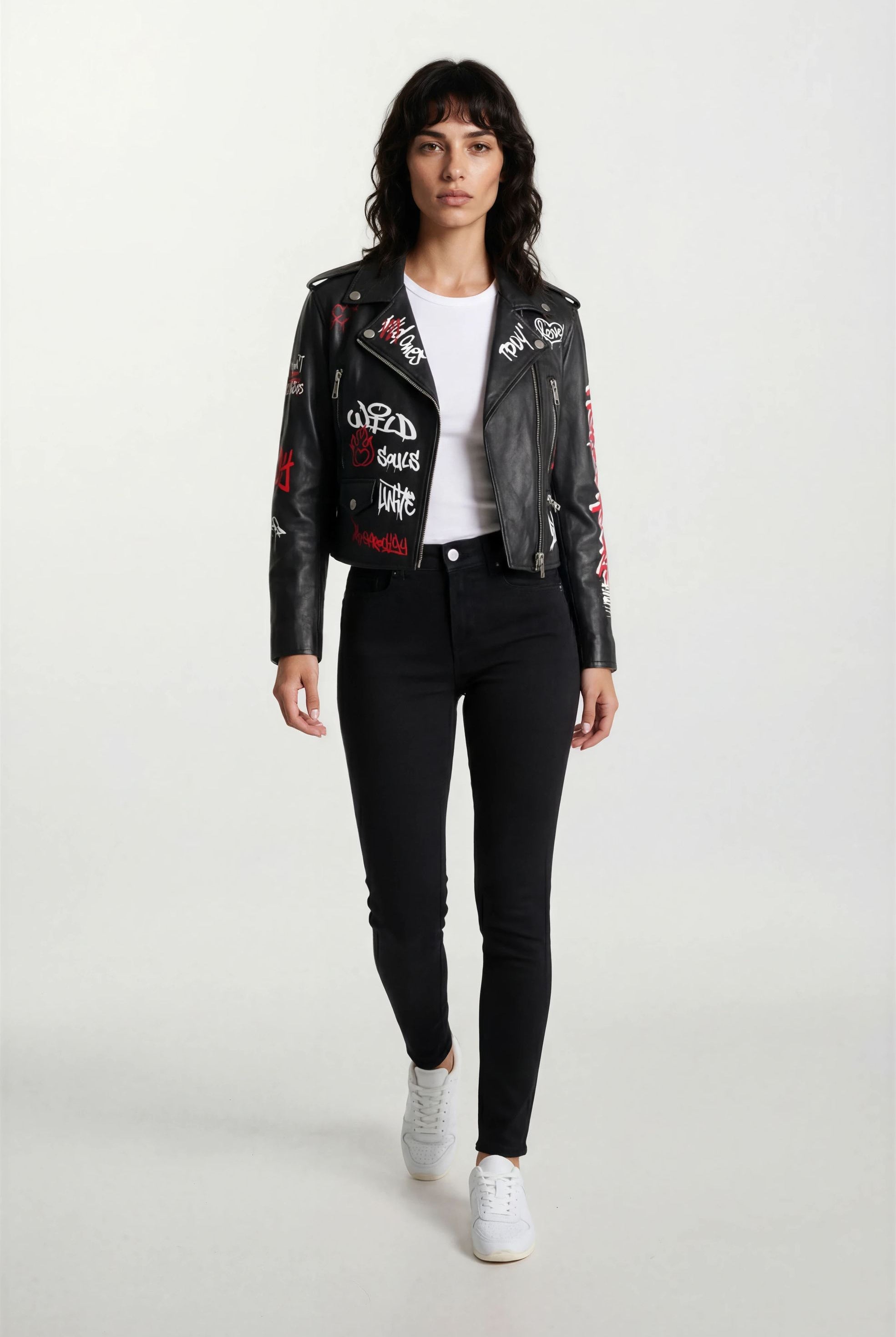 Eine Frau mit schulterlangen dunklen Haaren trägt die trueprodigy Feminine Lederjacke mit Grafittiprint und Reißverschluss - eine ausgefallene schwarze Lederjacke mit weißer und roter Grafik - mit einem weißen T-Shirt, schwarzen Jeans und weißen Turnschuhen und geht auf die Kamera zu.