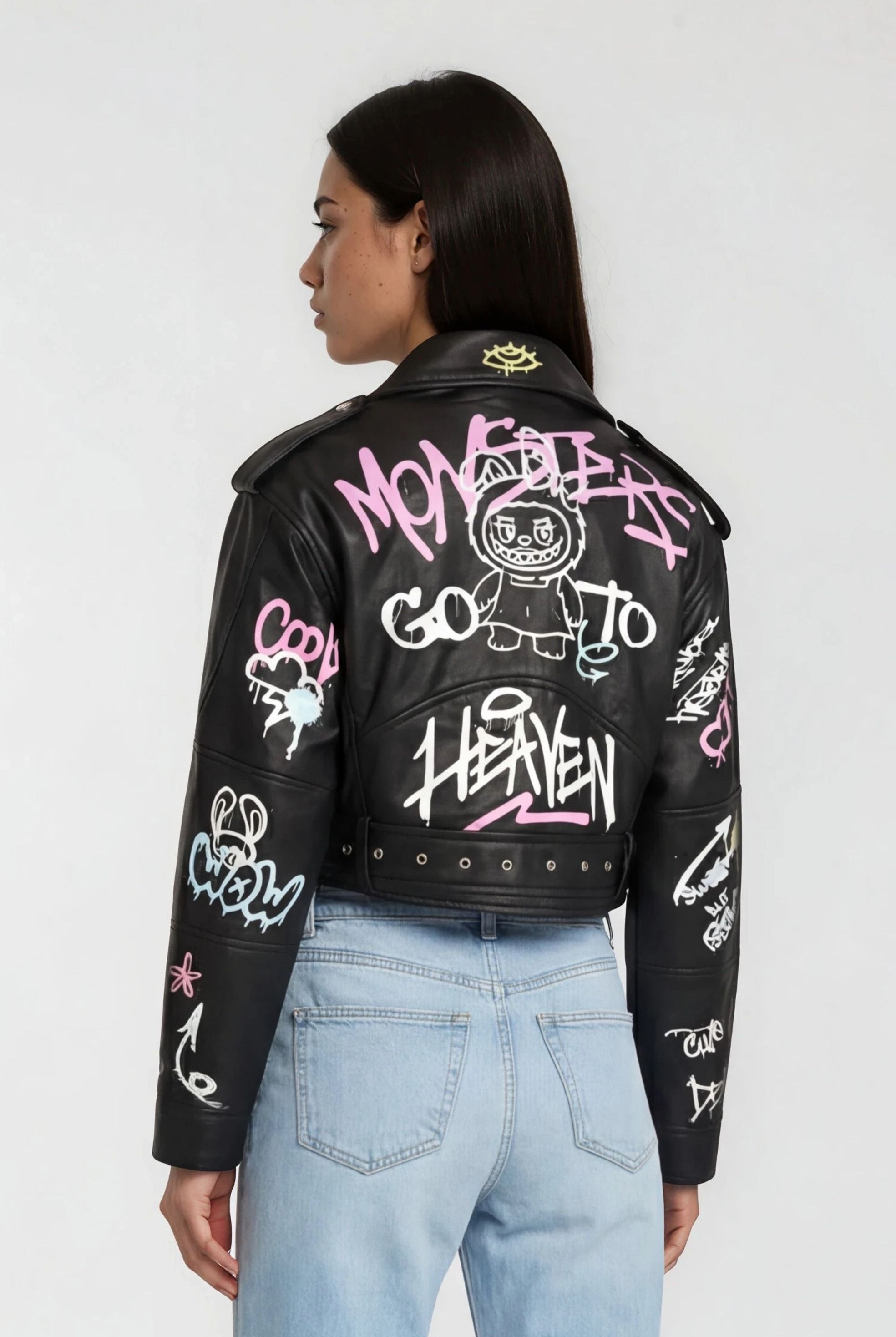 Eine Frau mit glattem dunklem Haar trägt eine trueprodigy Oversize Lederjacke mit Grafittiprint und Gürtel, auf der bunte Kunst und Texte wie HEAVEN und Cartoon-Gesichter zu sehen sind, gepaart mit hellblauen Jeans. Sie ist von hinten und von der Seite abgebildet.