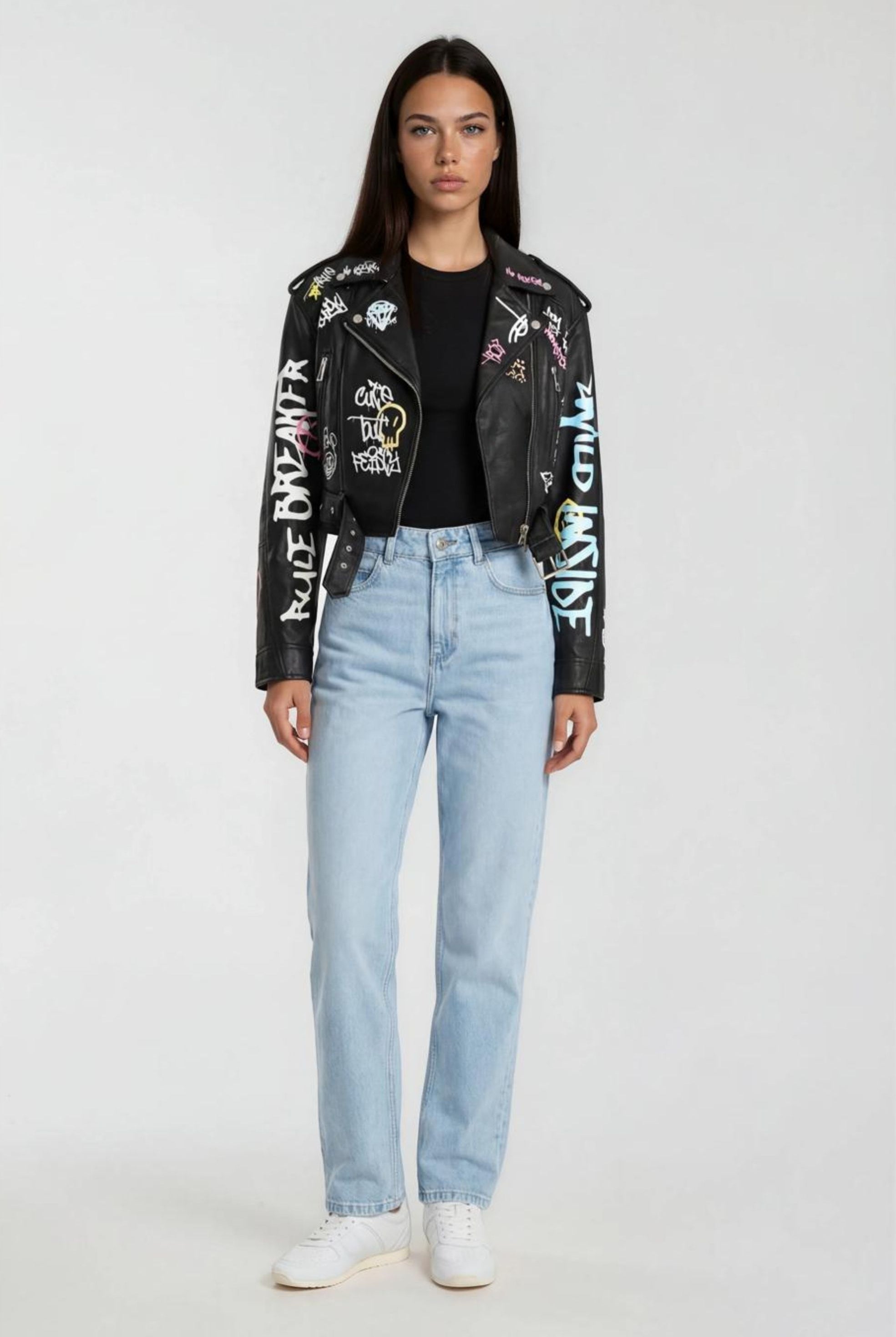 Eine Frau mit glattem dunklem Haar blickt nach vorne und trägt eine schwarze Oversize Lederjacke mit Grafittiprint und Gürtel von trueprodigy, ein schwarzes T-Shirt, hellblaue Jeans und weiße Turnschuhe vor einem schlichten hellen Hintergrund.