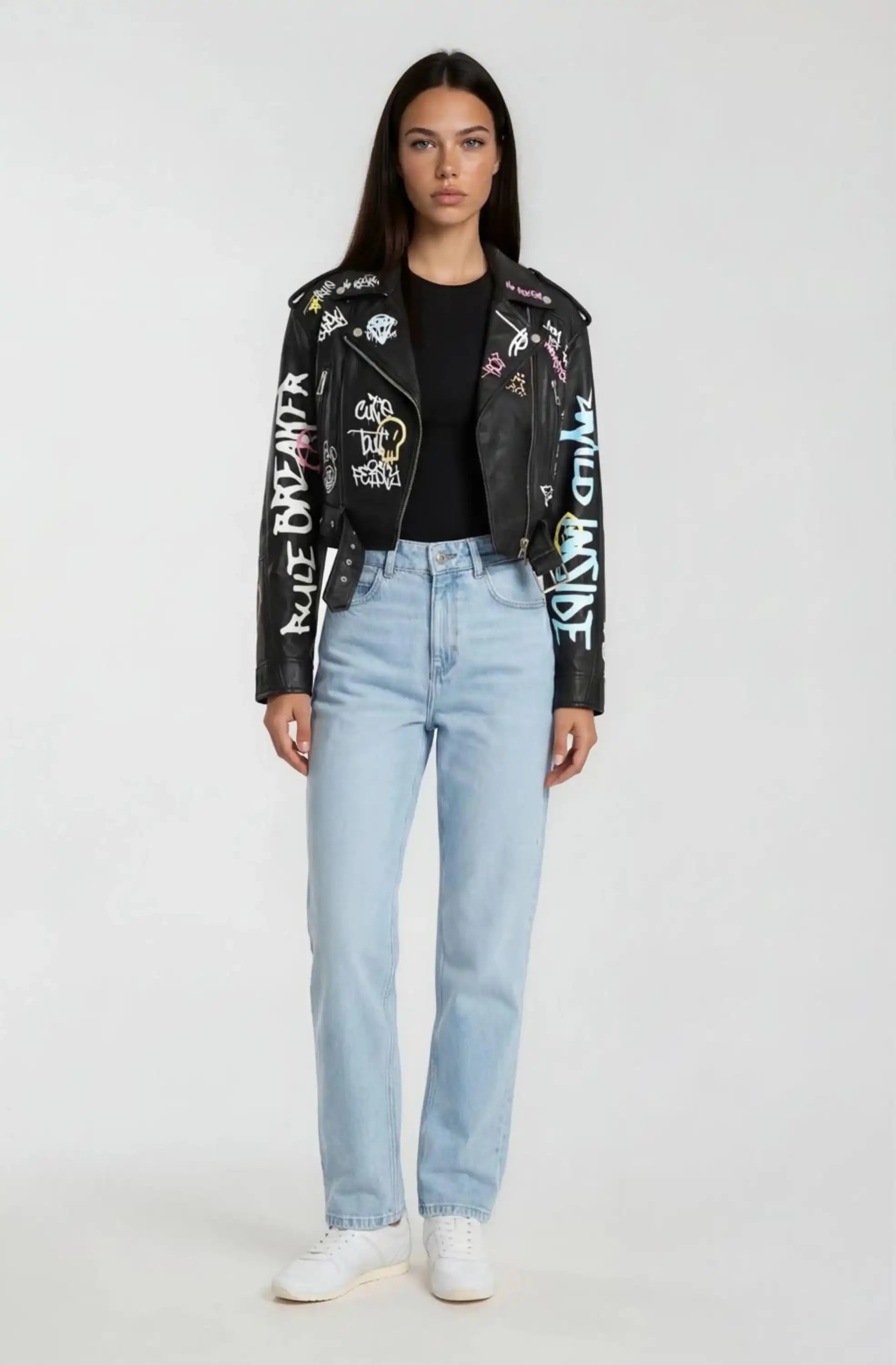 Eine Frau mit glattem dunklem Haar blickt nach vorne und trägt eine schwarze Oversize Lederjacke mit Grafittiprint und Gürtel von trueprodigy, ein schwarzes T-Shirt, hellblaue Jeans und weiße Turnschuhe vor einem schlichten hellen Hintergrund.