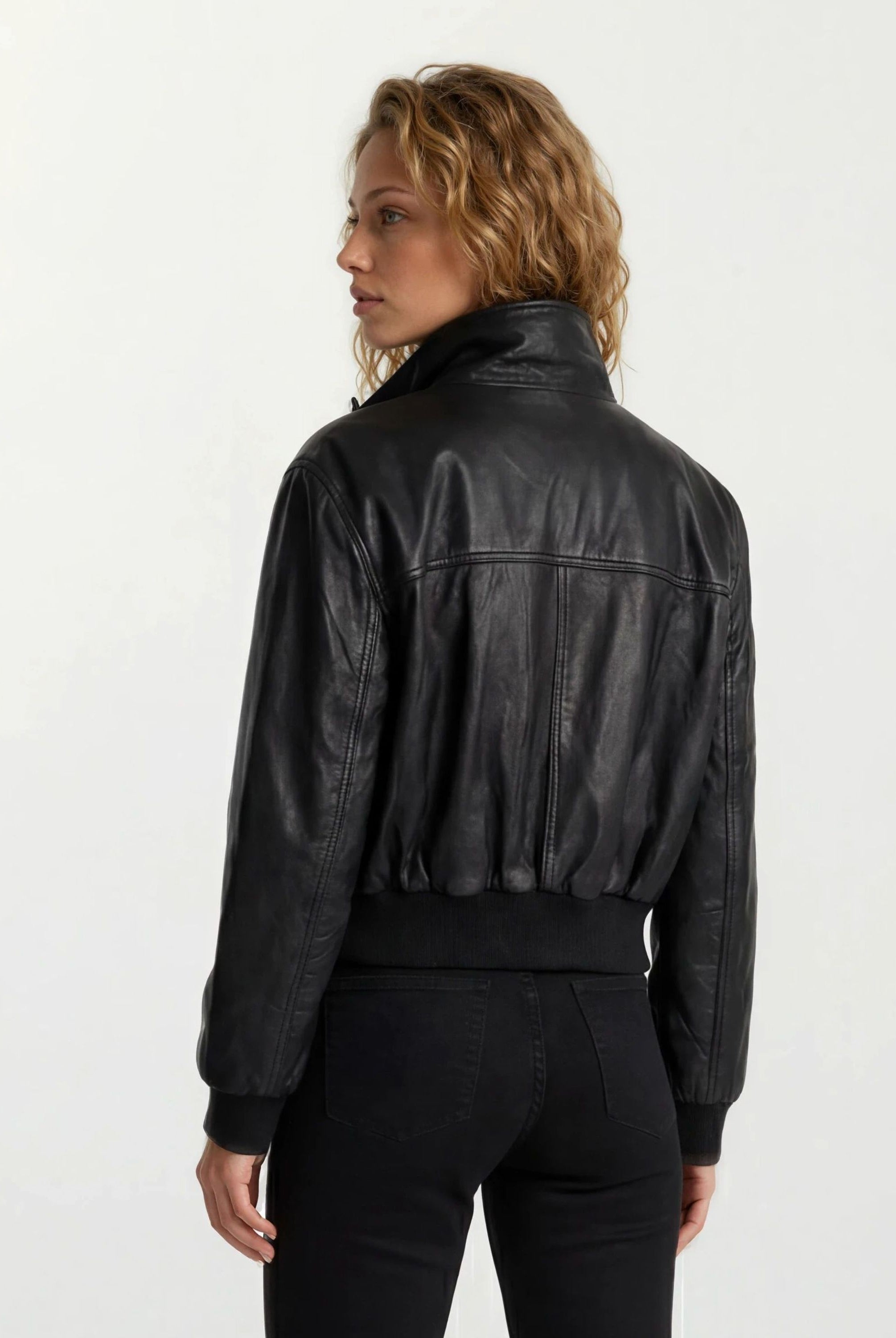 Abgebildet ist eine Person mit gewelltem, schulterlangem Haar in der Rücken- und Seitenansicht, die die trueprodigy Feminine Lederjacke Stehkragen und Reißverschluss und eine schwarze Jeans vor einem schlichten weißen Hintergrund trägt.