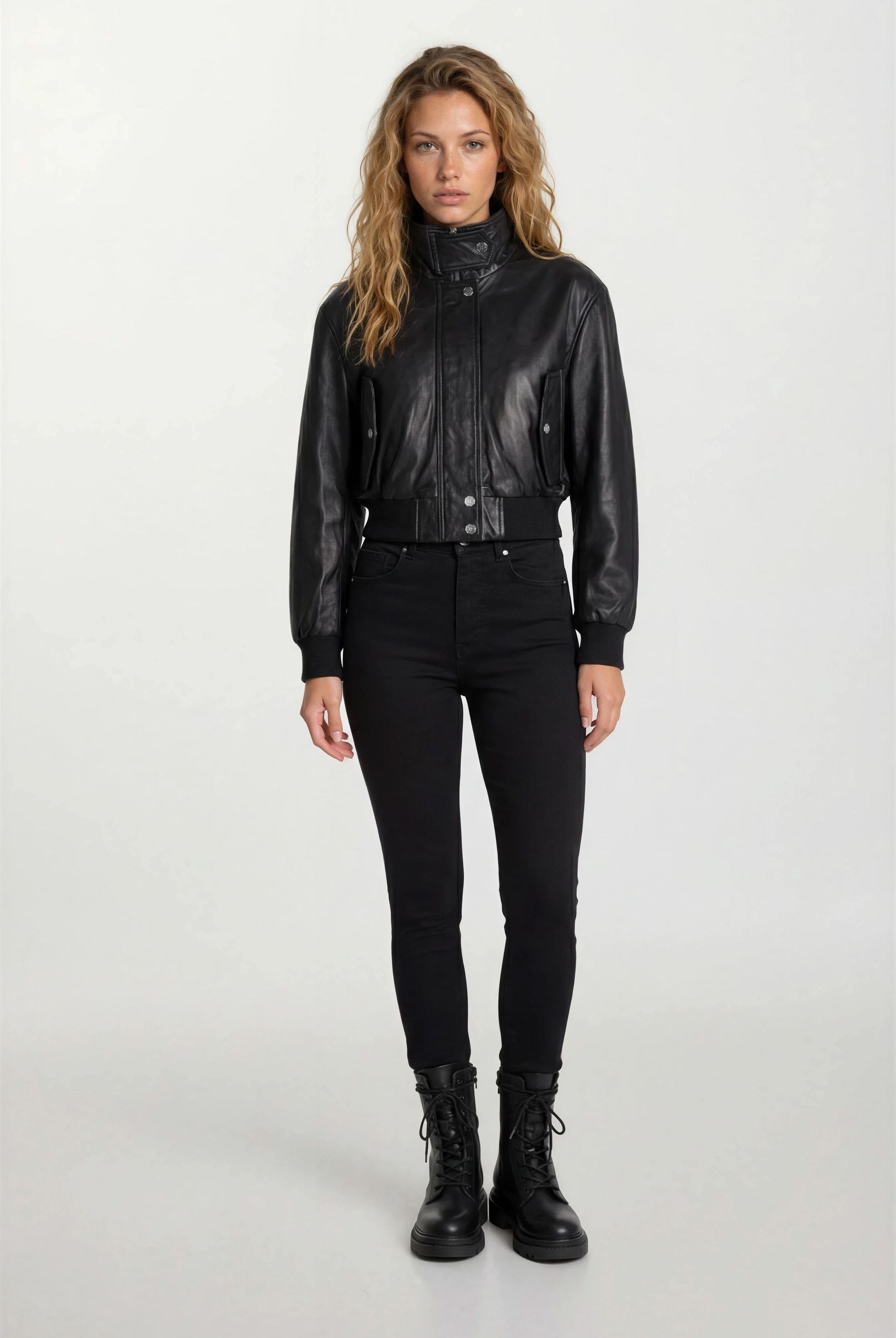 Eine Frau steht mit dem Gesicht nach vorne vor einem schlichten Hintergrund. Sie trägt die trueprodigy Feminine Lederjacke Stehkragen und Reißverschluss, eine schwarze Hose mit hoher Taille und schwarze Schnürstiefel. Sie hat die Hände an den Seiten und ihr langes, gewelltes Haar ist offen.