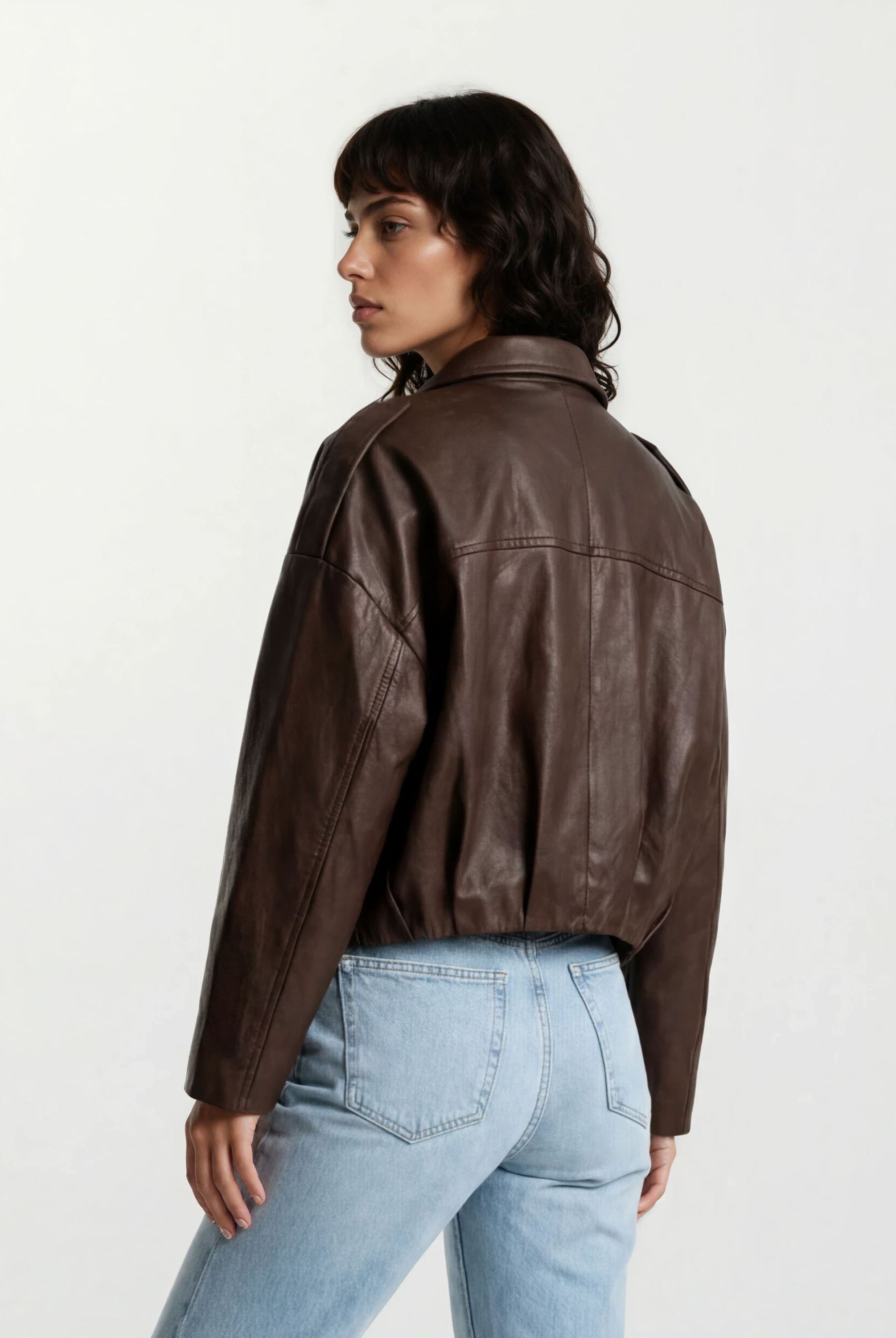 Eine Person mit gewellten dunklen Haaren trägt die trueprodigy Feminine Lederjacke mit Hemdkragen und Raffung am Saum und hellblaue Jeans und steht mit leicht gedrehtem Rücken und Kopf vor einem schlichten hellen Hintergrund.