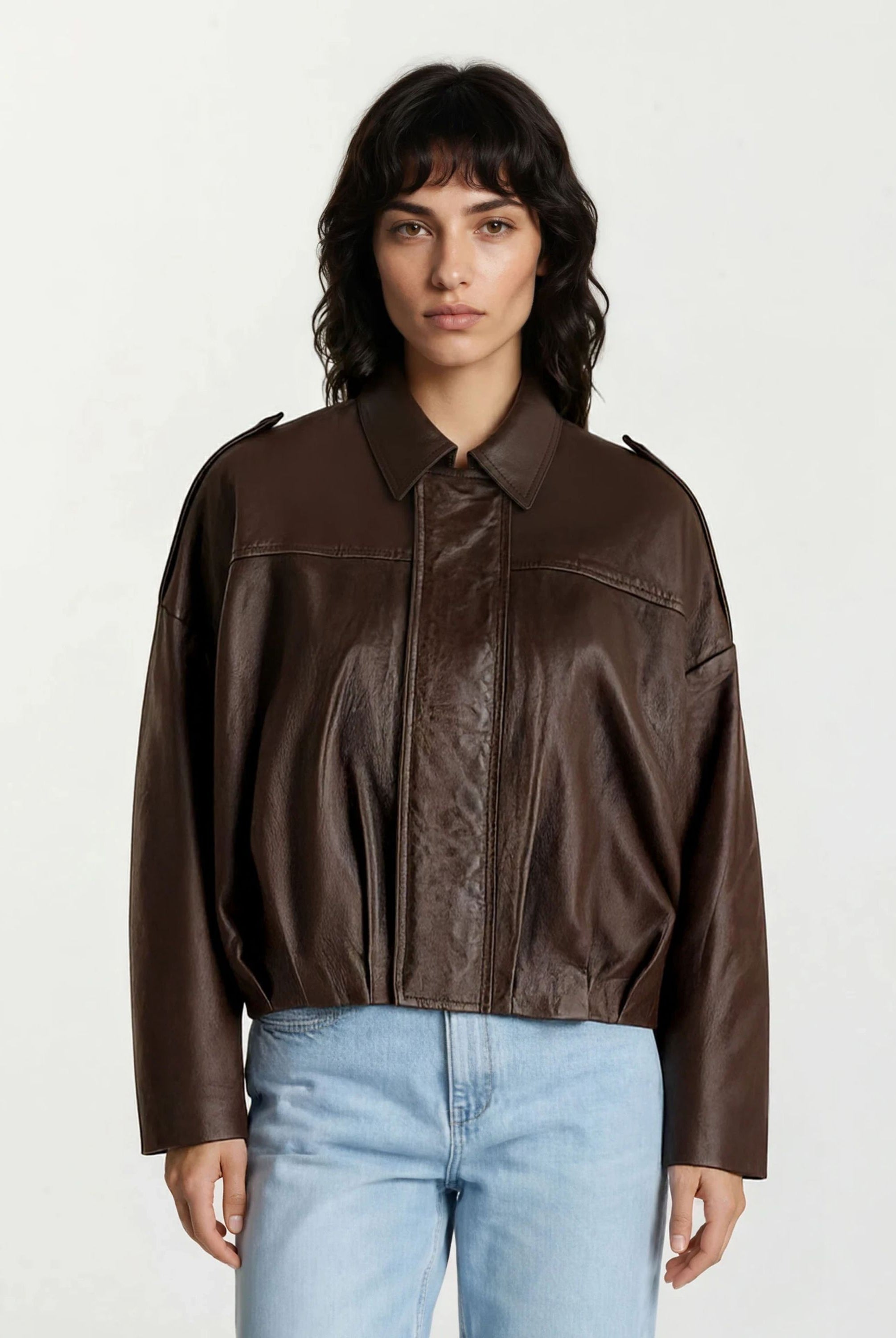 Eine Person mit schulterlangen dunklen Haaren und Pony modelliert die trueprodigy Feminine Lederjacke mit Hemdkragen und Raffung am Saum in Dunkelbraun, gepaart mit hellblauen Jeans, vor einem schlichten, hellen Hintergrund.