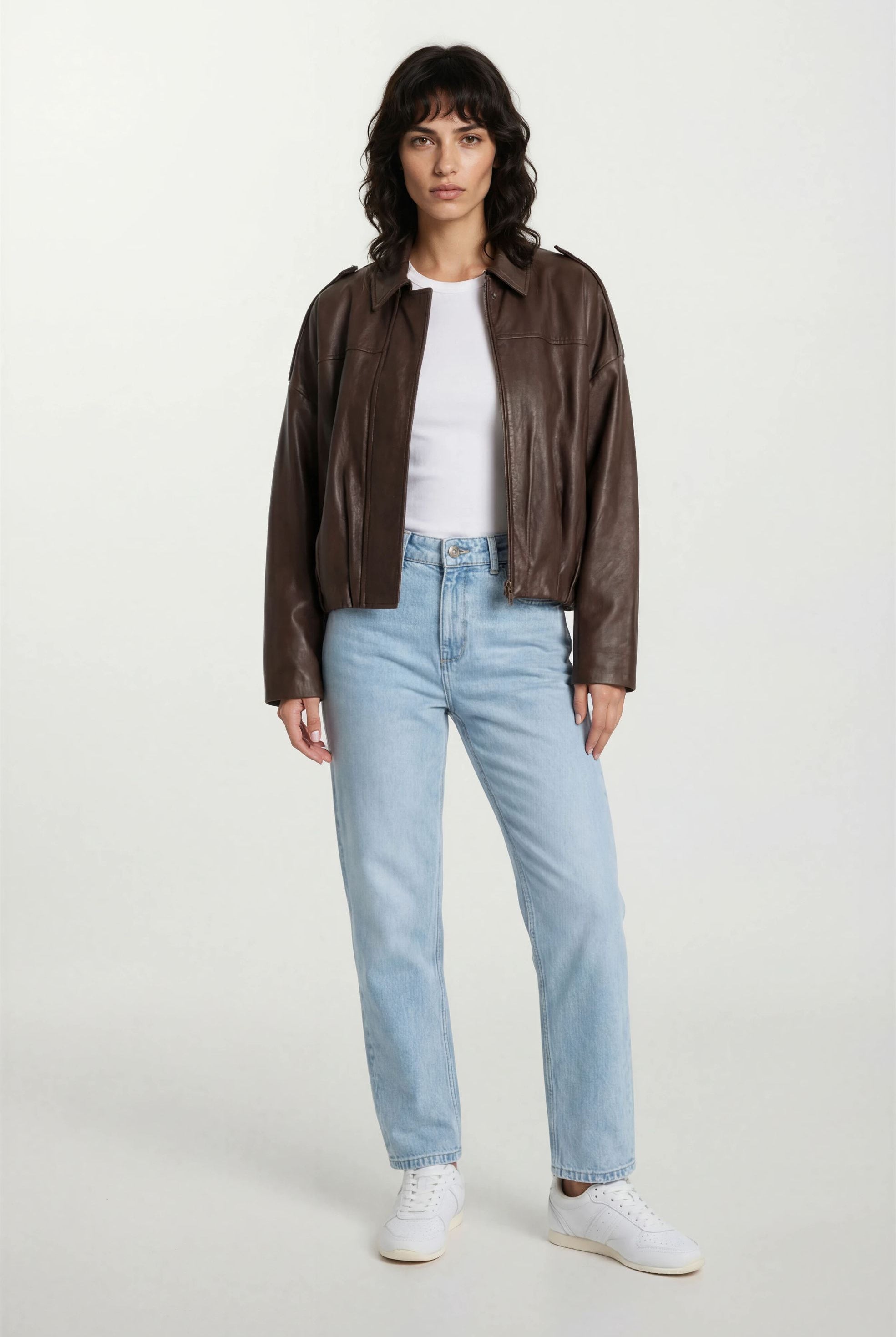 Eine Person trägt eine wahrhaftig feminine Lederjacke mit Hemdkragen und Raffung am Saum, dazu ein weißes T-Shirt, hellblaue Jeans und weiße Sneaker vor einem schlichten, hellen Hintergrund.