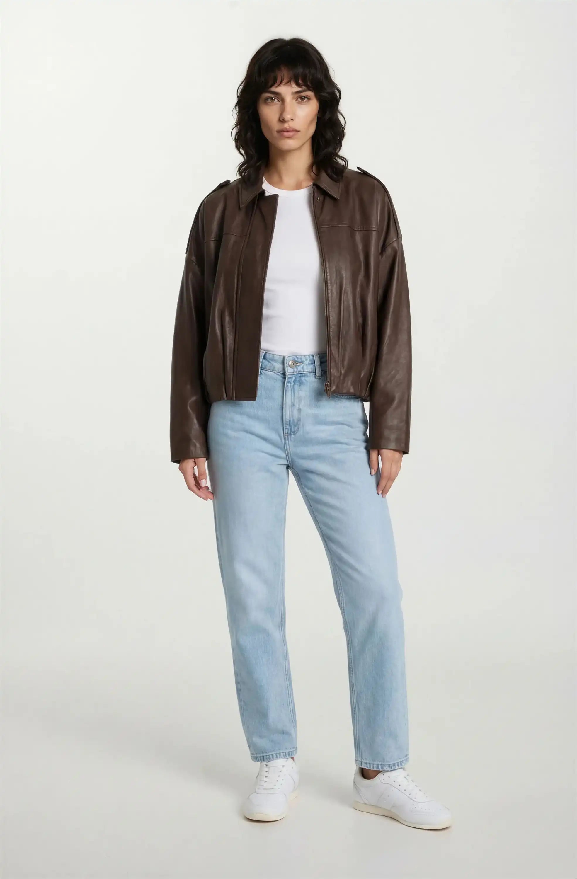 Eine Person trägt eine wahrhaftig feminine Lederjacke mit Hemdkragen und Raffung am Saum, dazu ein weißes T-Shirt, hellblaue Jeans und weiße Sneaker vor einem schlichten, hellen Hintergrund.