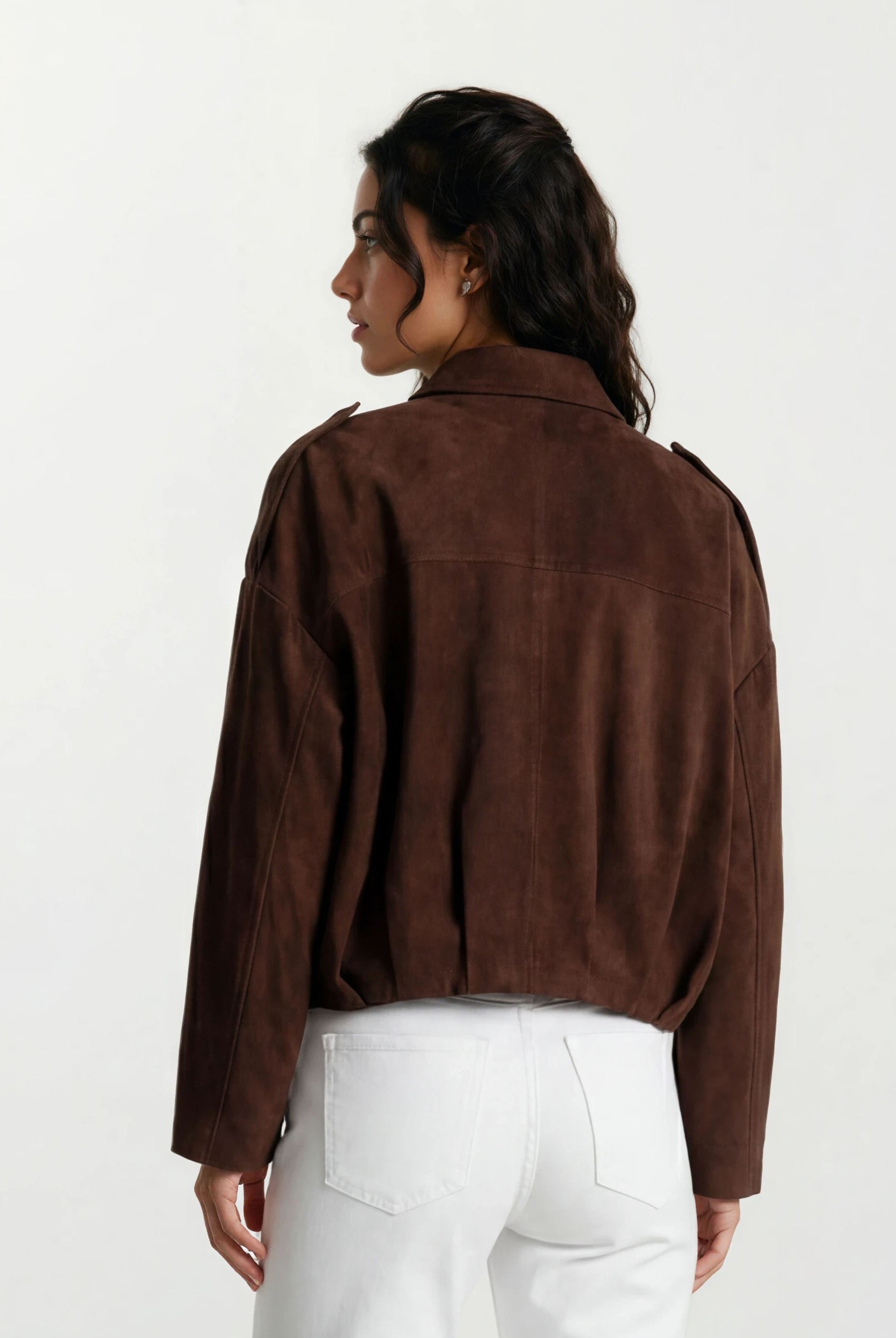 Eine Frau mit dunklem gewelltem Haar, von hinten gesehen, trägt die trueprodigy Feminine Lederjacke mit Hemdkragen und Raffung am Saum in brauner und weißer Hose vor einem schlichten, hellen Hintergrund.