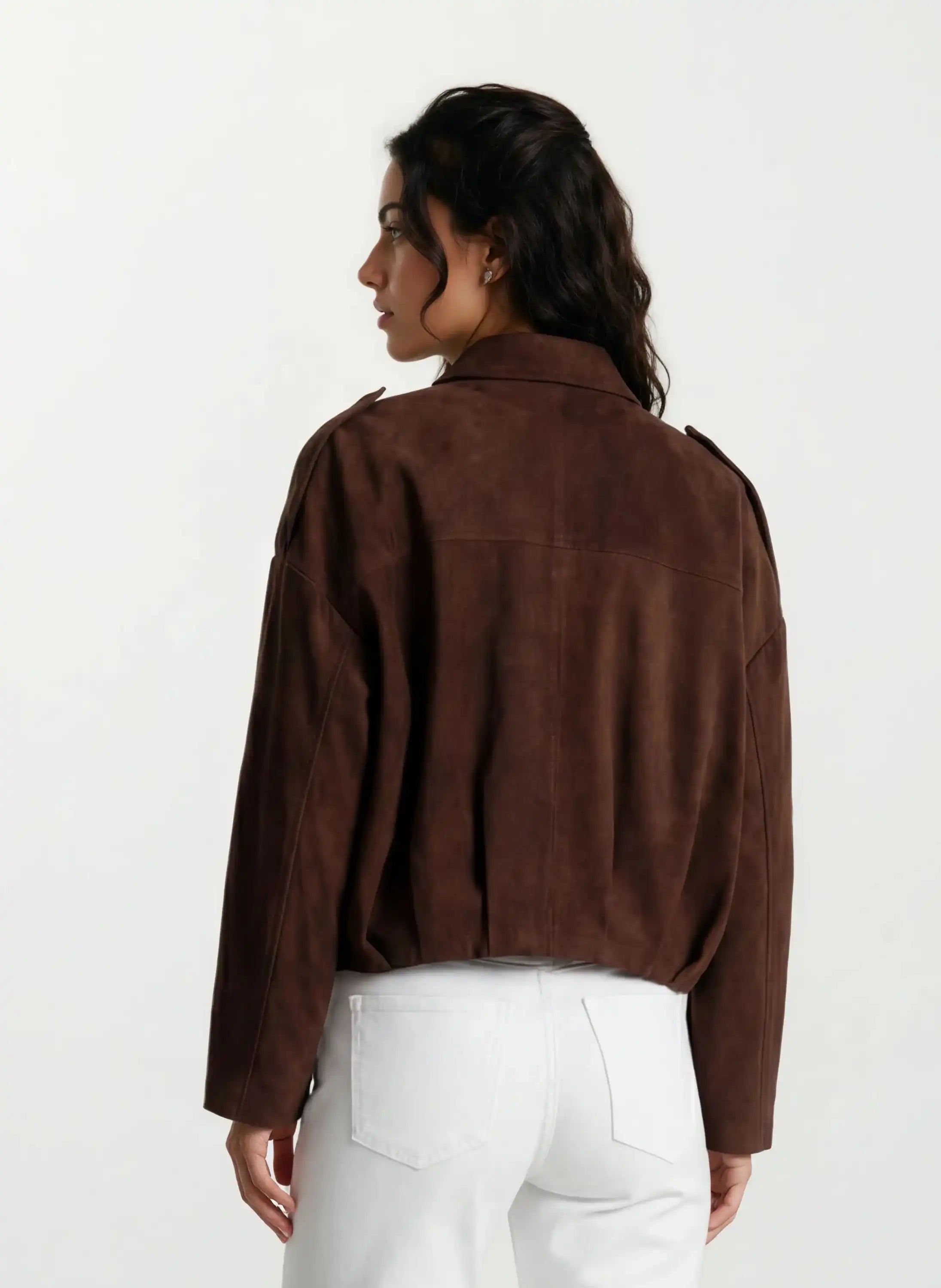 Eine Frau mit dunklem gewelltem Haar, von hinten gesehen, trägt die trueprodigy Feminine Lederjacke mit Hemdkragen und Raffung am Saum in brauner und weißer Hose vor einem schlichten, hellen Hintergrund.