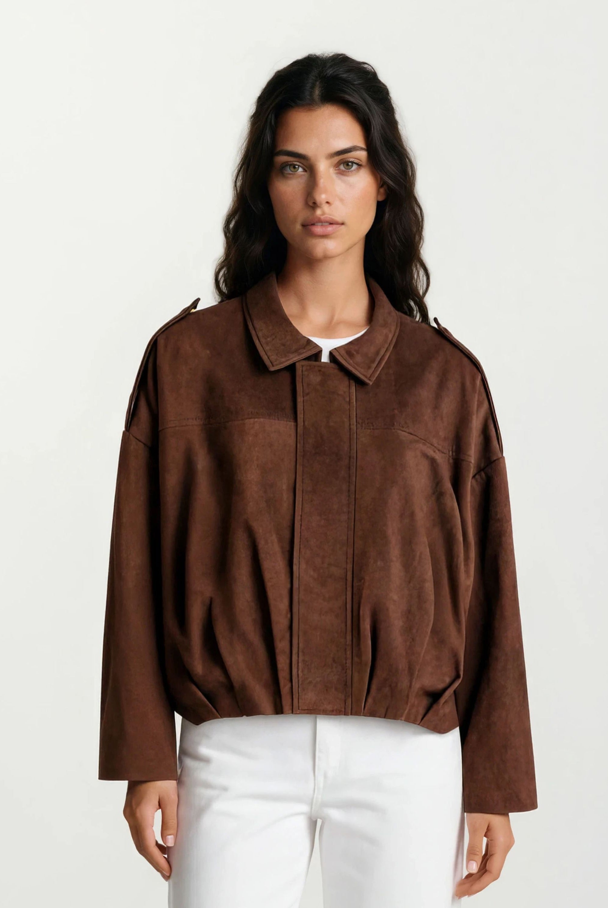 Eine Person mit langen dunklen Haaren trägt die trueprodigy Feminine Lederjacke mit Hemdkragen und Raffung am Saum und eine weiße Hose, die vor einem schlichten hellen Hintergrund steht.