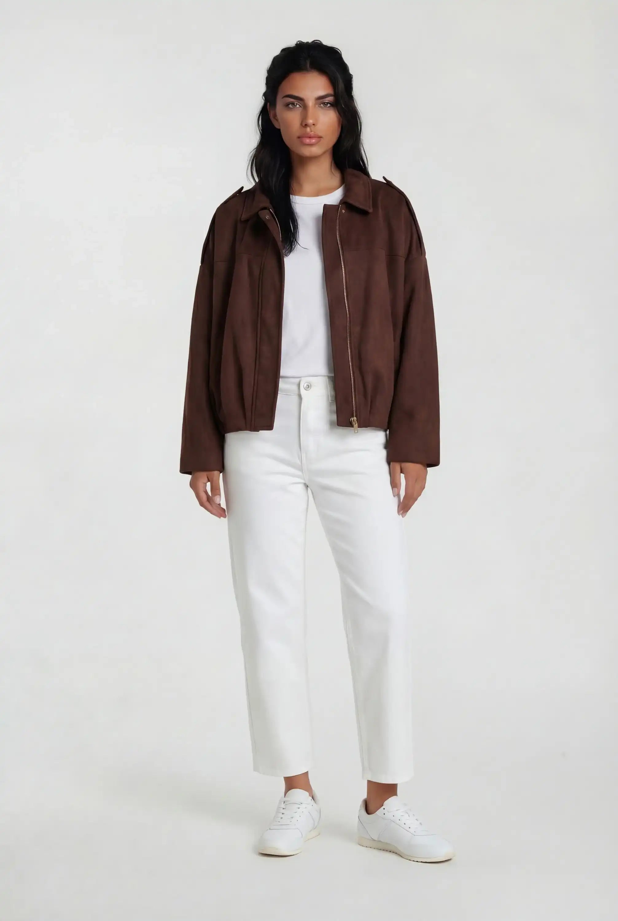 Eine Person steht mit dem Gesicht nach vorne und trägt die trueprodigy Clean-Cut Wildlederjacke Damen "Naeko" mit Hemdkragen, ein weißes T-Shirt, eine weiße Hose mit hoher Taille und weiße Turnschuhe vor einem schlichten hellen Hintergrund.