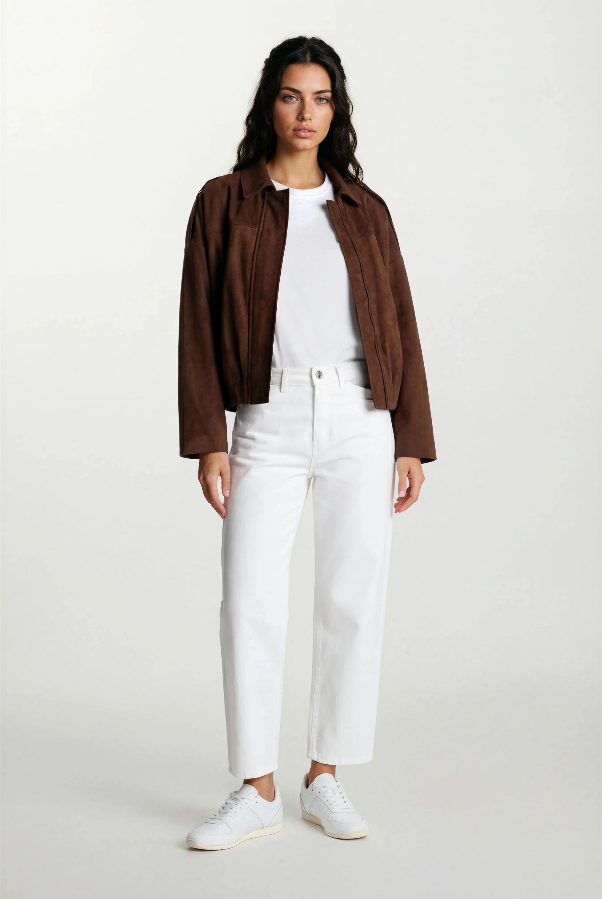 Eine Frau mit langen dunklen Haaren und neutralem Gesichtsausdruck trägt die trueprodigy Feminine Lederjacke mit Hemdkragen und Raffung am Saum über einem weißen T-Shirt, einer weißen Hose mit hoher Taille und weißen Turnschuhen vor einem schlichten hellen Hintergrund.