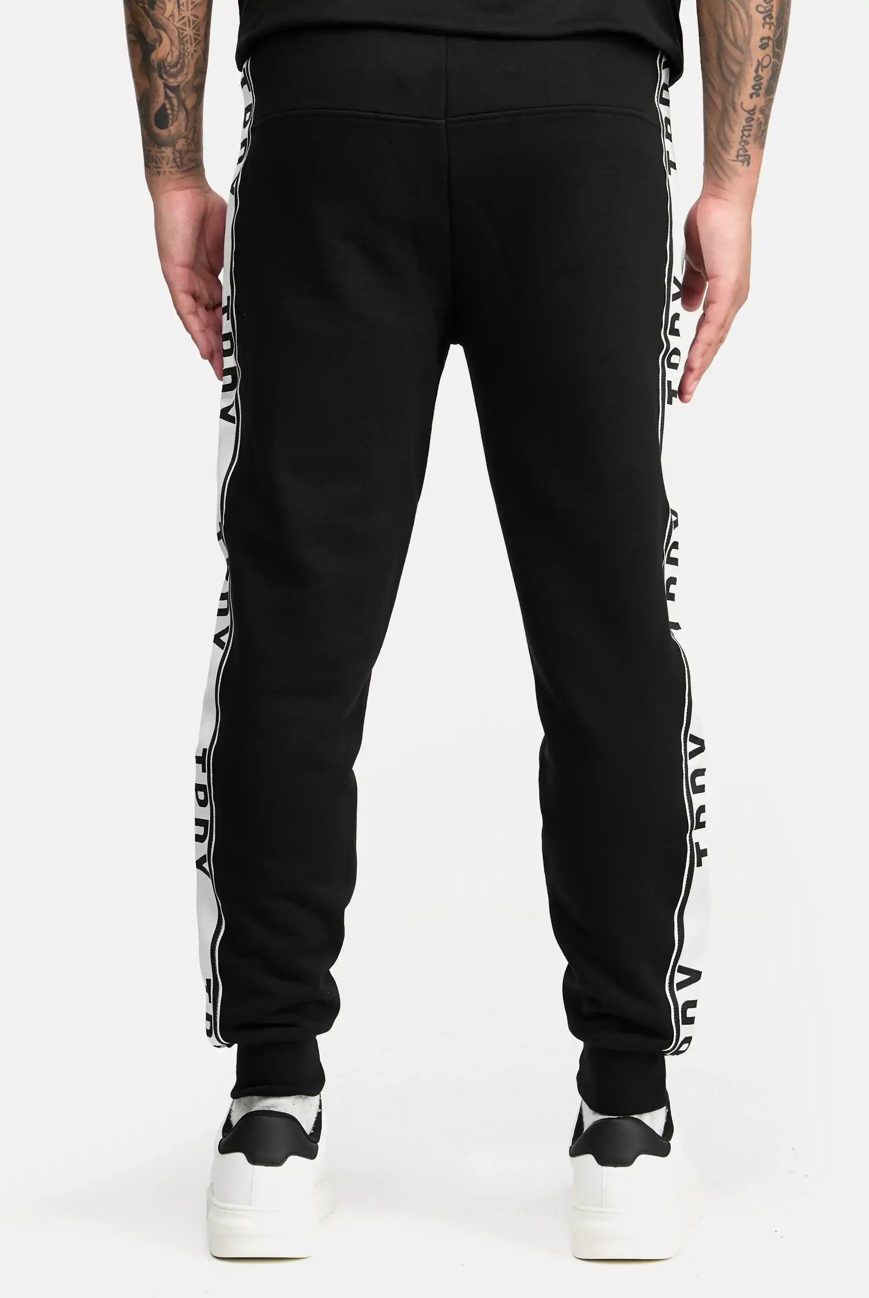 Eine Person trägt die trueprodigy Weston Sweathose mit Logoschrift. Die stylische Hose im Regular Fit mit Kontraststreifen passt perfekt zu weißen Sneakern und Tattoos vor schlichtem, weißem Hintergrund.