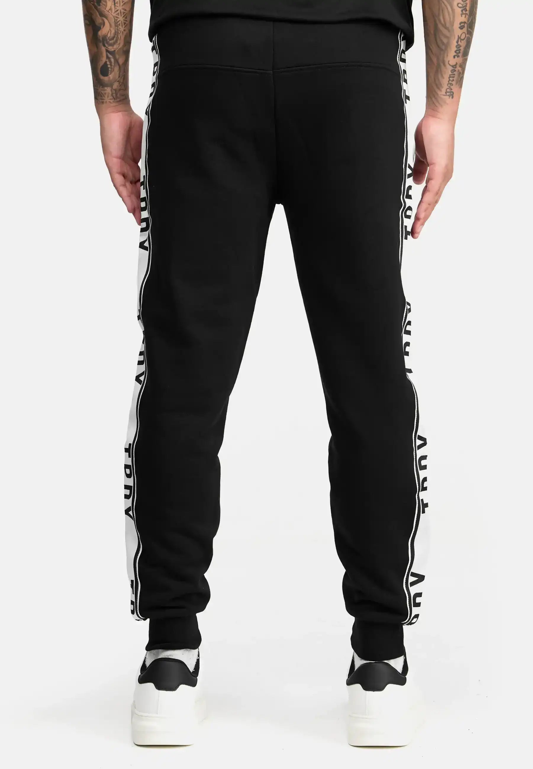 Eine Person trägt die trueprodigy Weston Sweathose mit Logoschrift. Die stylische Hose im Regular Fit mit Kontraststreifen passt perfekt zu weißen Sneakern und Tattoos vor schlichtem, weißem Hintergrund.