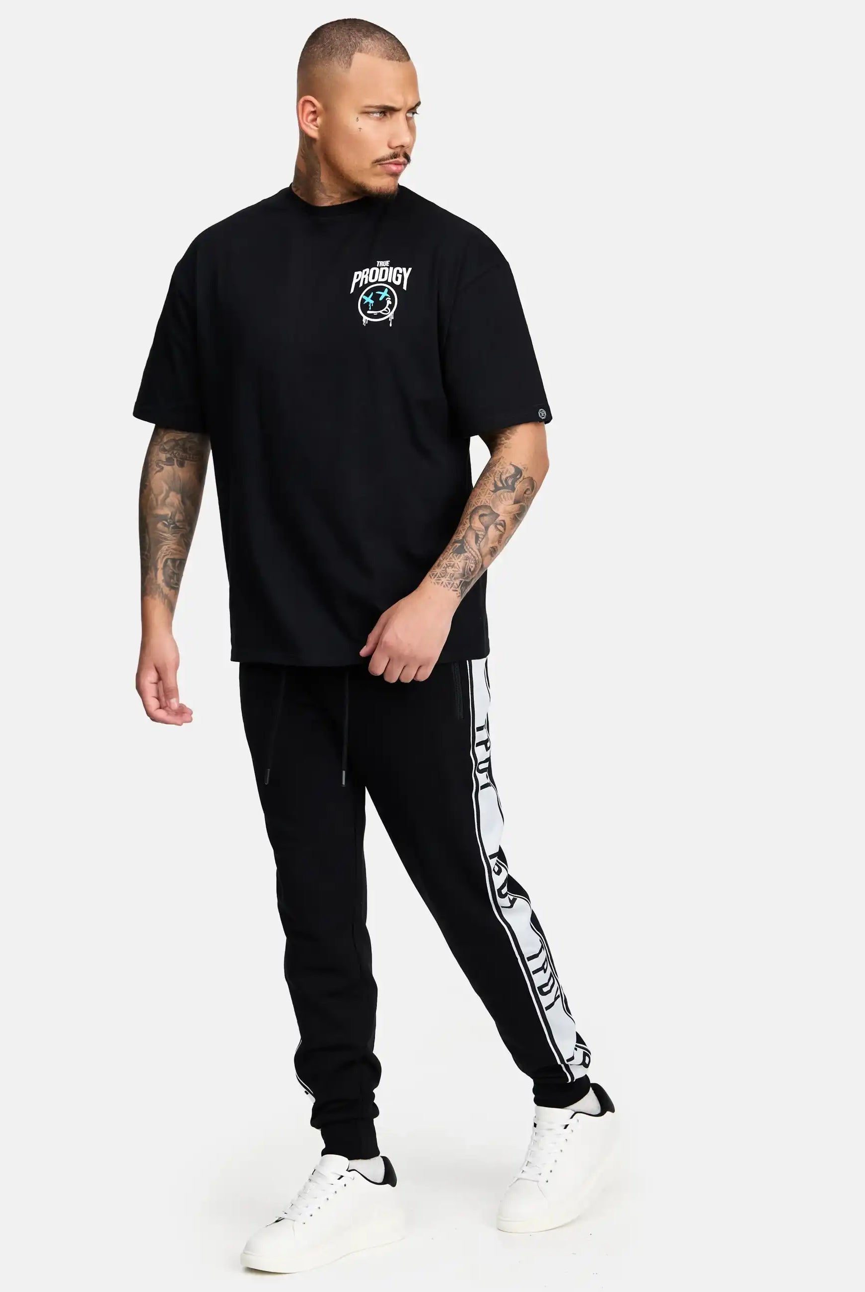 Eine Person mit kurzen Haaren und Tattoos trägt ein schwarzes Hemd mit PRODIGY-Text und -Grafik, Weston Sweathose mit Logoschrift von trueprodigy, und weiße Turnschuhe und steht vor einem schlichten hellen Hintergrund.