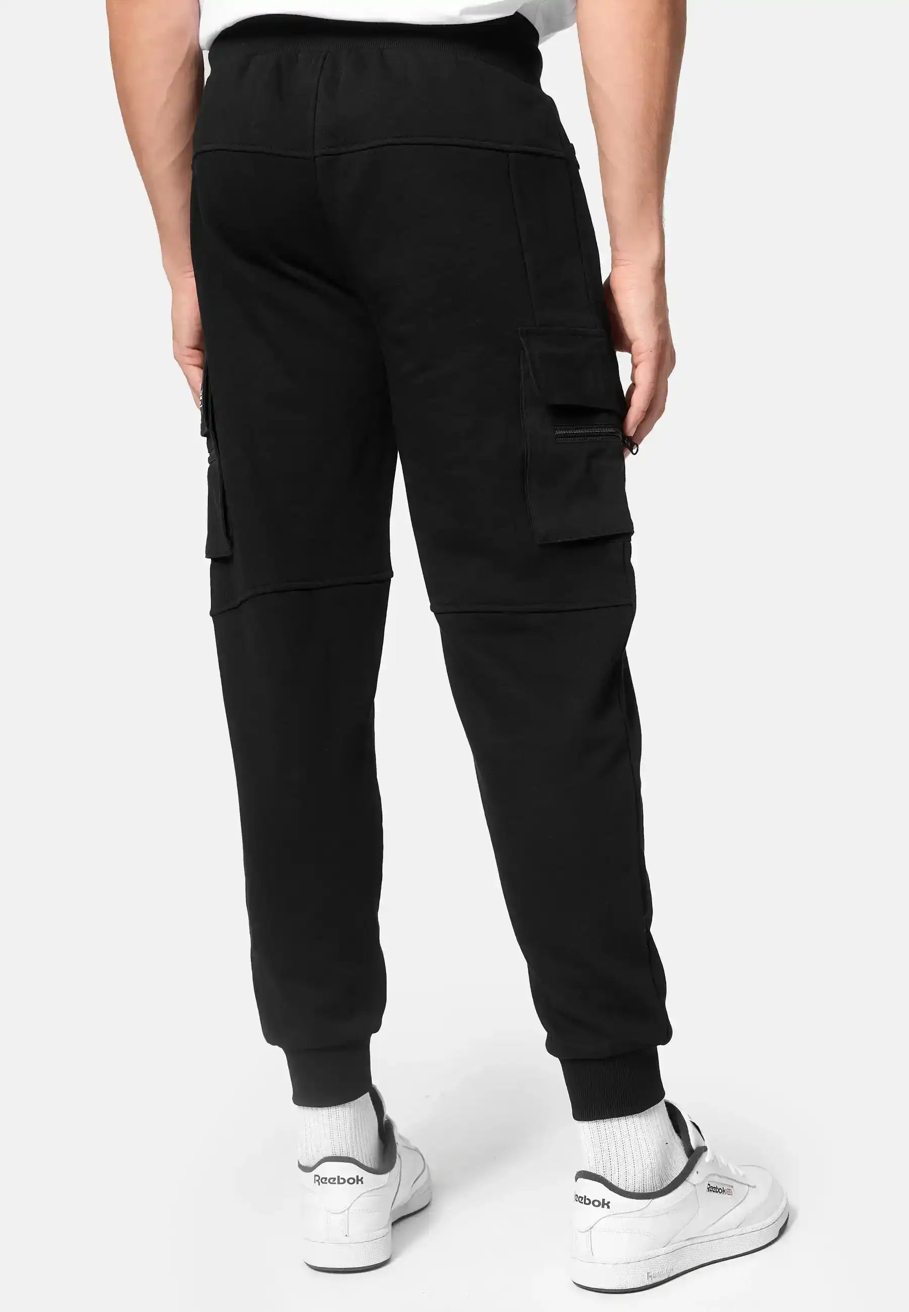 Eine Person steht mit dem Rücken zur Kamera und trägt trueprodigy George Cargo Sweathose in Schwarz, gepaart mit weißen Socken und weißen Reebok Turnschuhen vor einem schlichten Hintergrund.