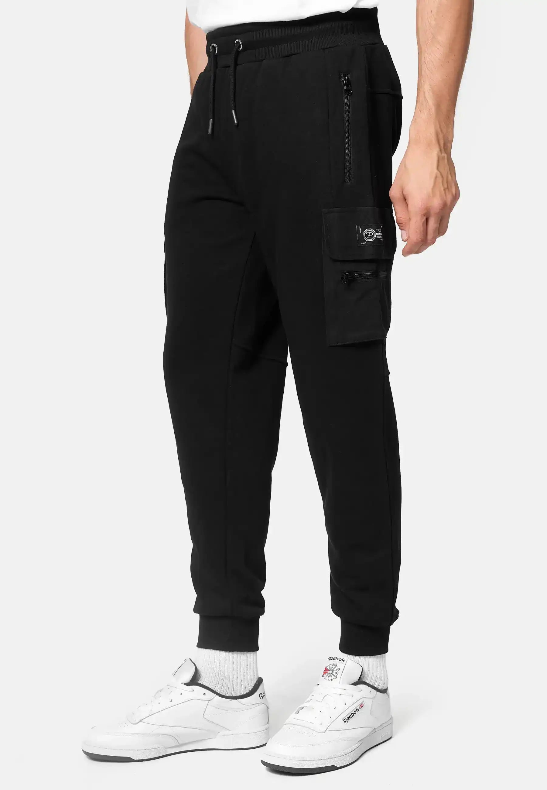 Eine Person trägt eine schwarze George Cargo Sweathose von trueprodigy mit Reißverschluss und Pattentaschen, weiße Socken und weiße Turnschuhe vor einem schlichten hellen Hintergrund. Nur die untere Hälfte ist zu sehen - idealer Cargohose Herren-Stil.