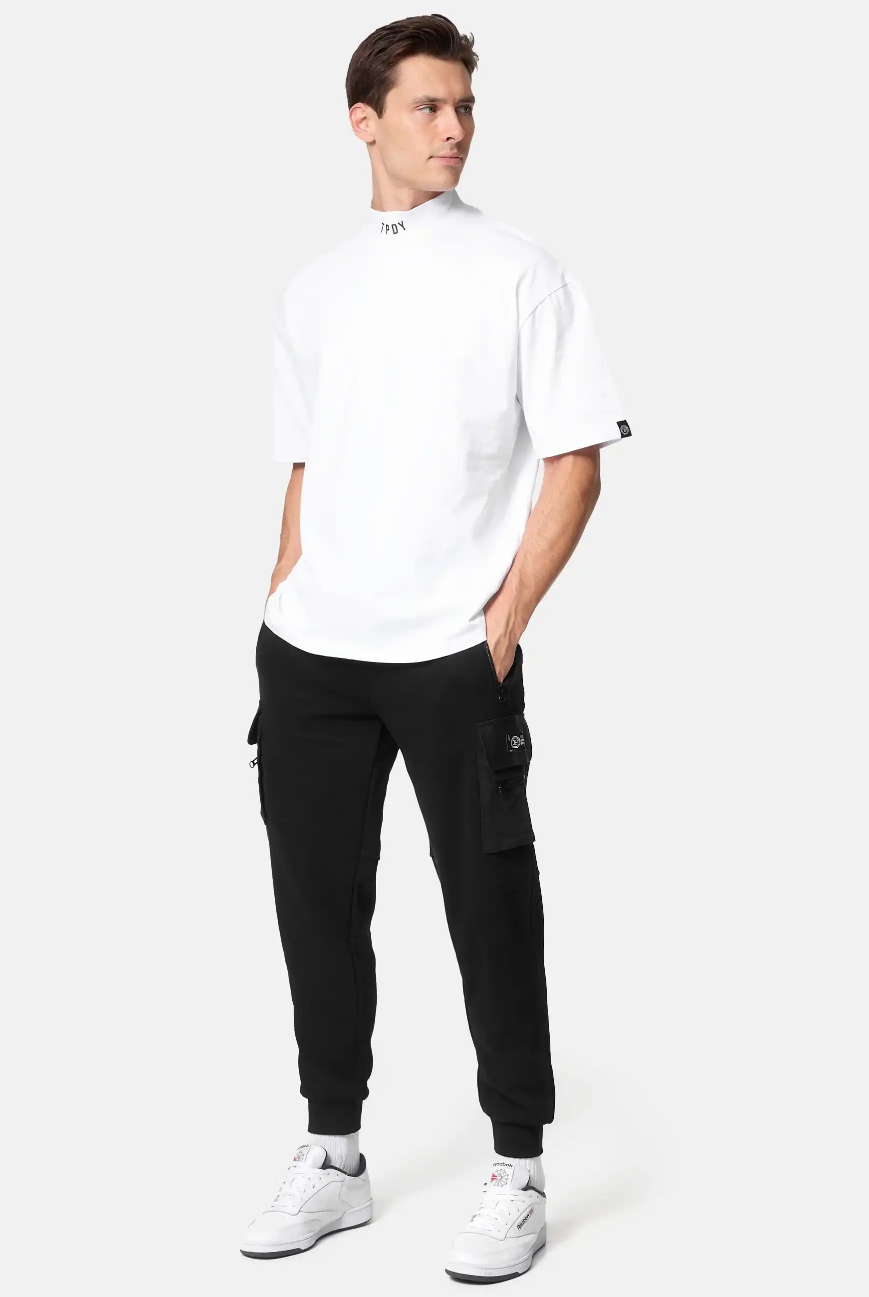 Eine Person steht mit den Händen in den Taschen, trägt ein weißes Oversized-T-Shirt, trueprodigy George Cargo Sweathose in Schwarz und weiße Turnschuhe vor einem schlichten hellgrauen Hintergrund.