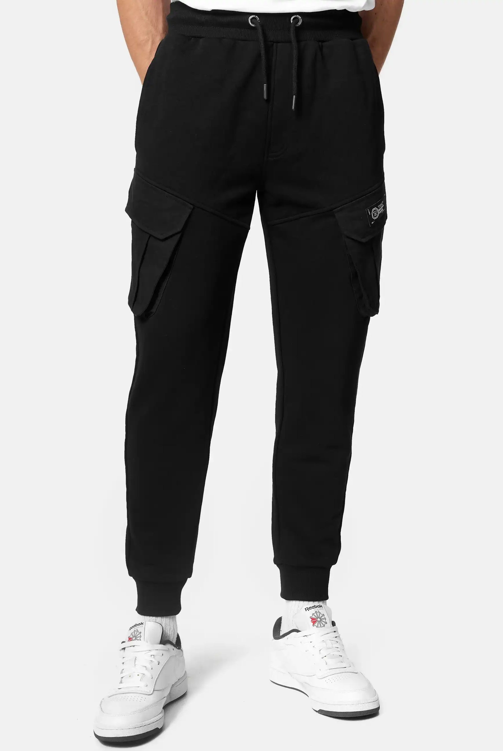 Eine Person in schwarzer Stan Sweathose mit Cargotaschen von trueprodigy mit Seitentaschen und weißen Sneakern vor einem schlichten Hintergrund.