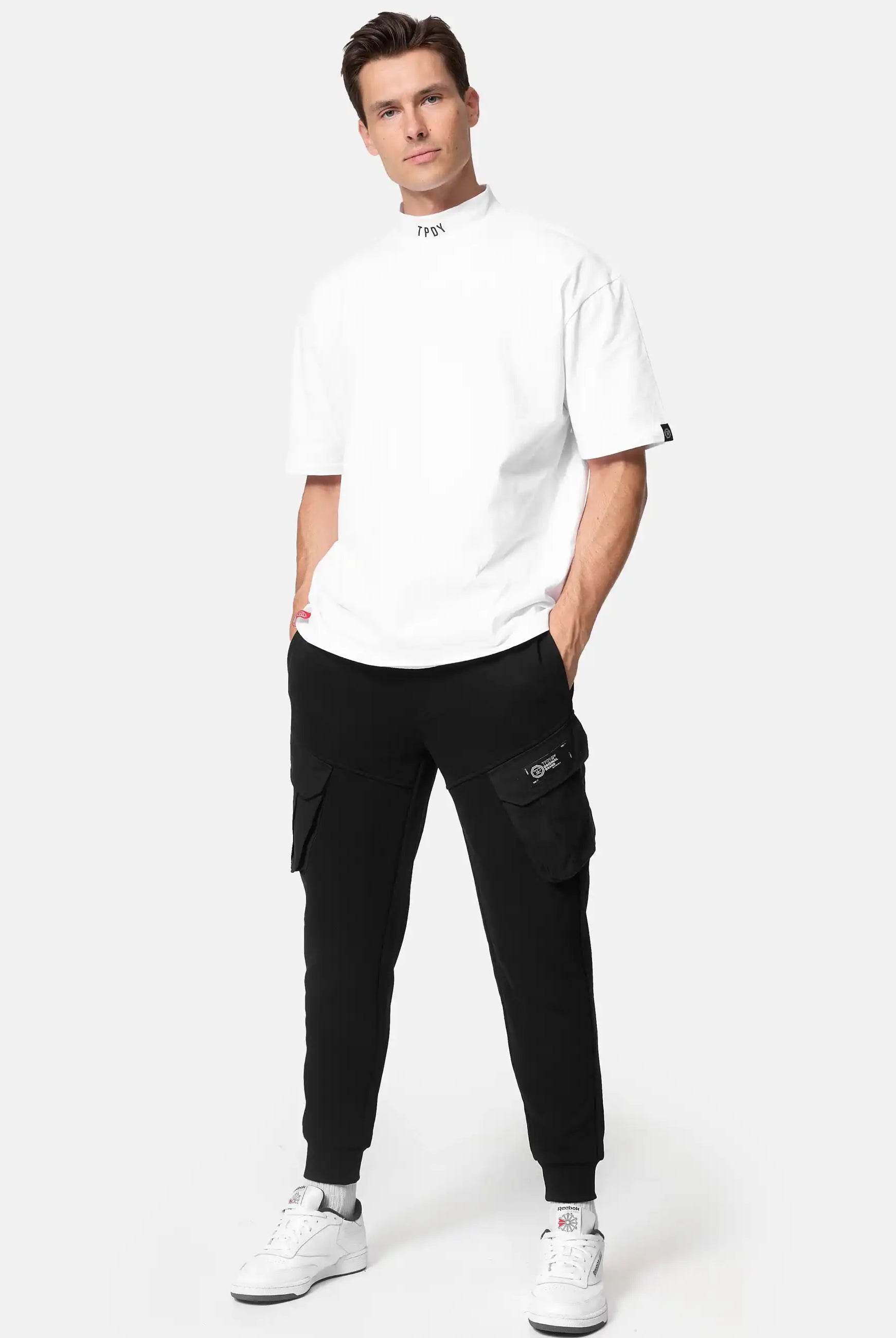 Ein Mann steht in einem weißen Oversized-T-Shirt, einer schwarzen trueprodigy Stan Sweathose mit Cargotaschen Jogginghose und weißen Turnschuhen vor einem schlichten hellen Hintergrund.