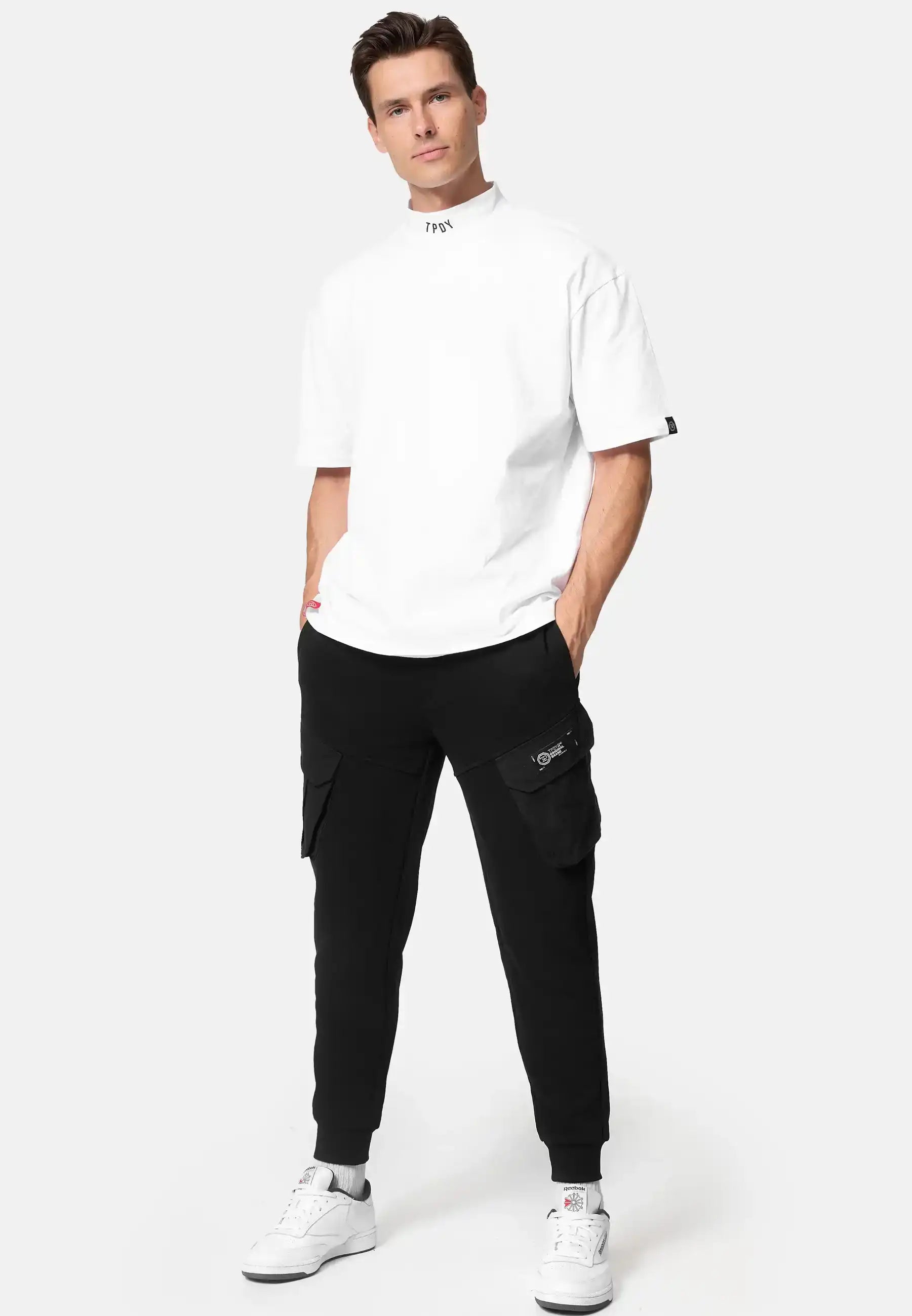 Ein Mann steht in einem weißen Oversized-T-Shirt, einer schwarzen trueprodigy Stan Sweathose mit Cargotaschen Jogginghose und weißen Turnschuhen vor einem schlichten hellen Hintergrund.