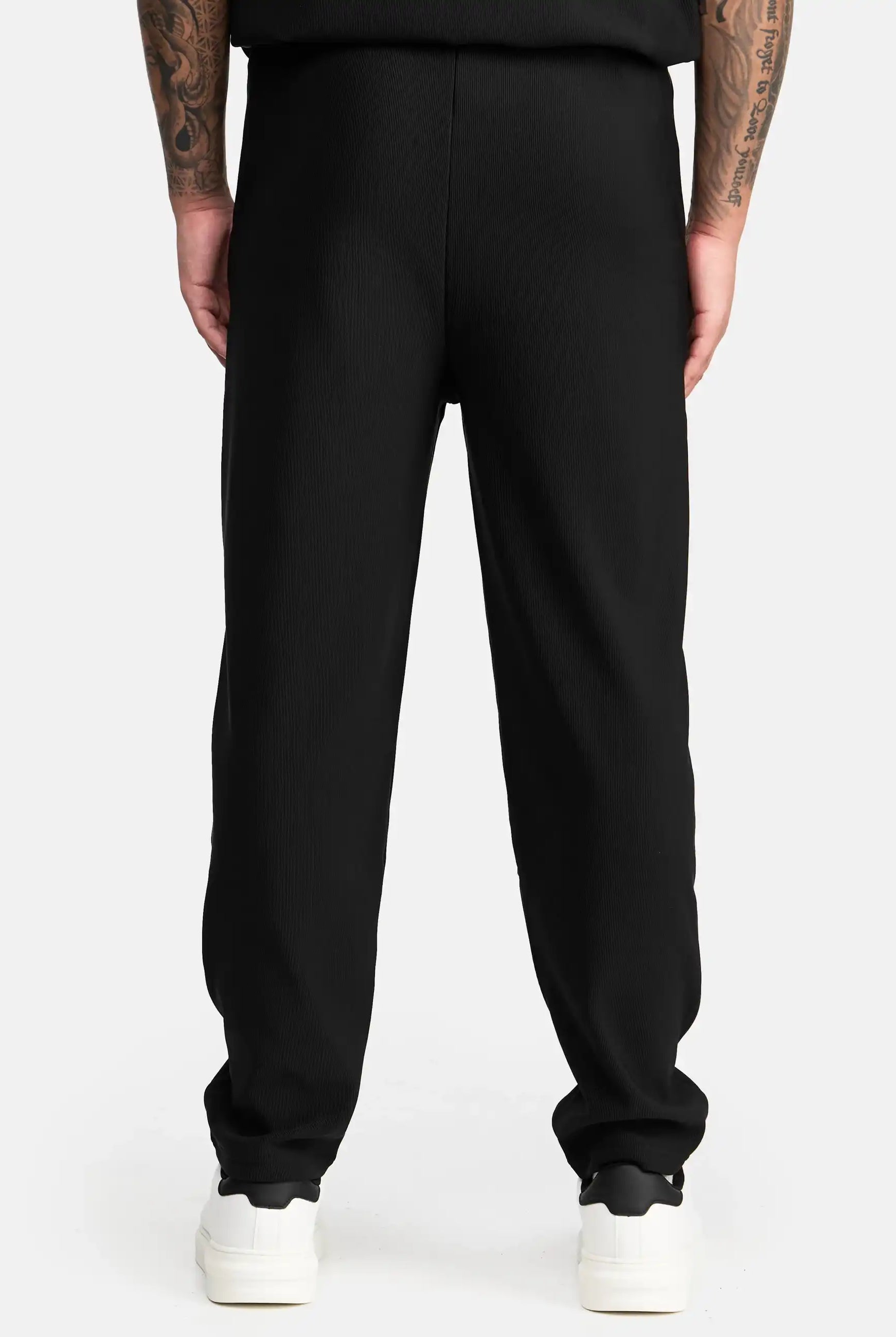 Eine Person steht mit dem Rücken zur Kamera und trägt trueprodigy Ewan Jogger mit Stickerei in schwarzen und weißen Turnschuhen. Auf ihren Armen, die seitlich herabhängen, sind Tätowierungen zu sehen. Der Hintergrund ist einfarbig und hell gefärbt.