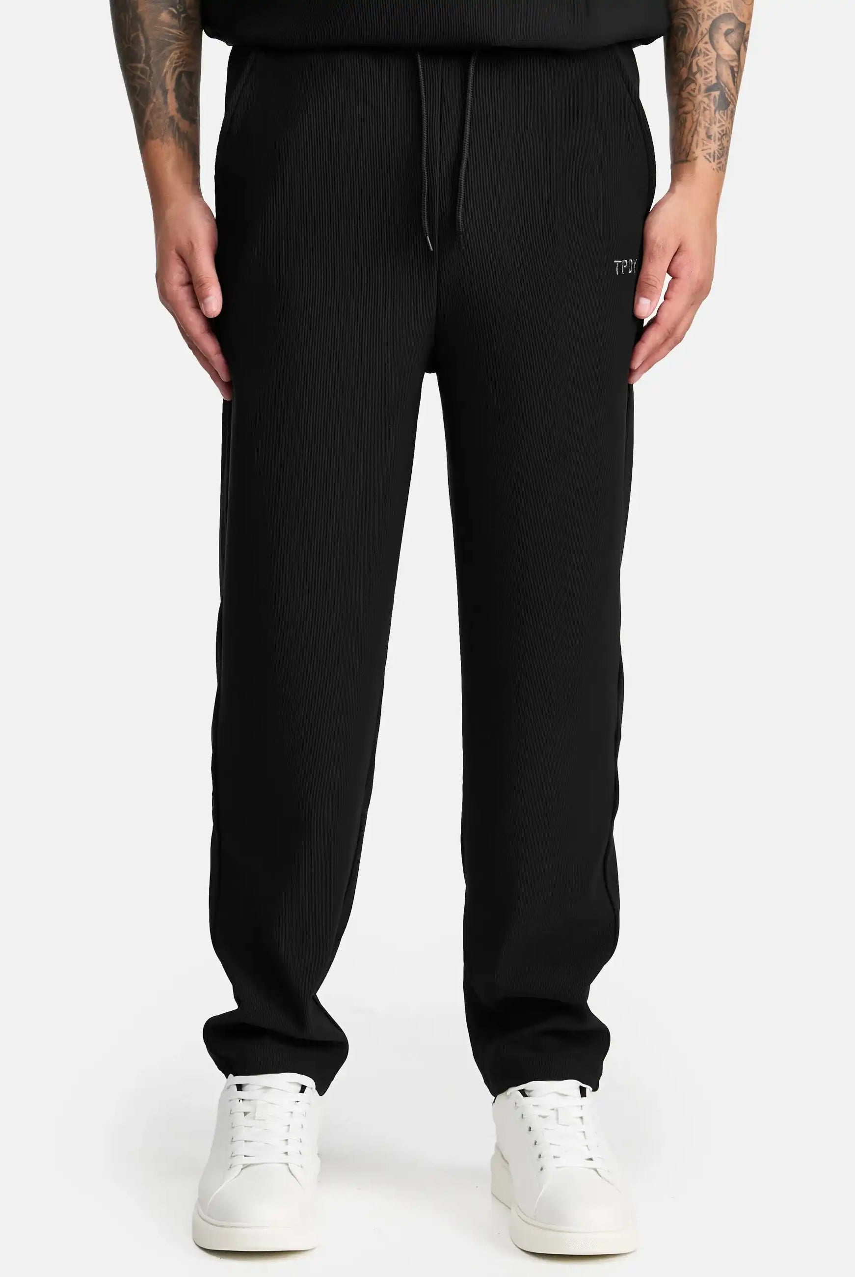 Eine Person trägt den trueprodigy Ewan Jogger mit Stickerei mit Kordelzug in der Taille, weiße Turnschuhe und ein schwarzes Oberteil. Nur der Unterkörper ist sichtbar und zeigt trueprodigy Tattoos auf beiden Unterarmen.