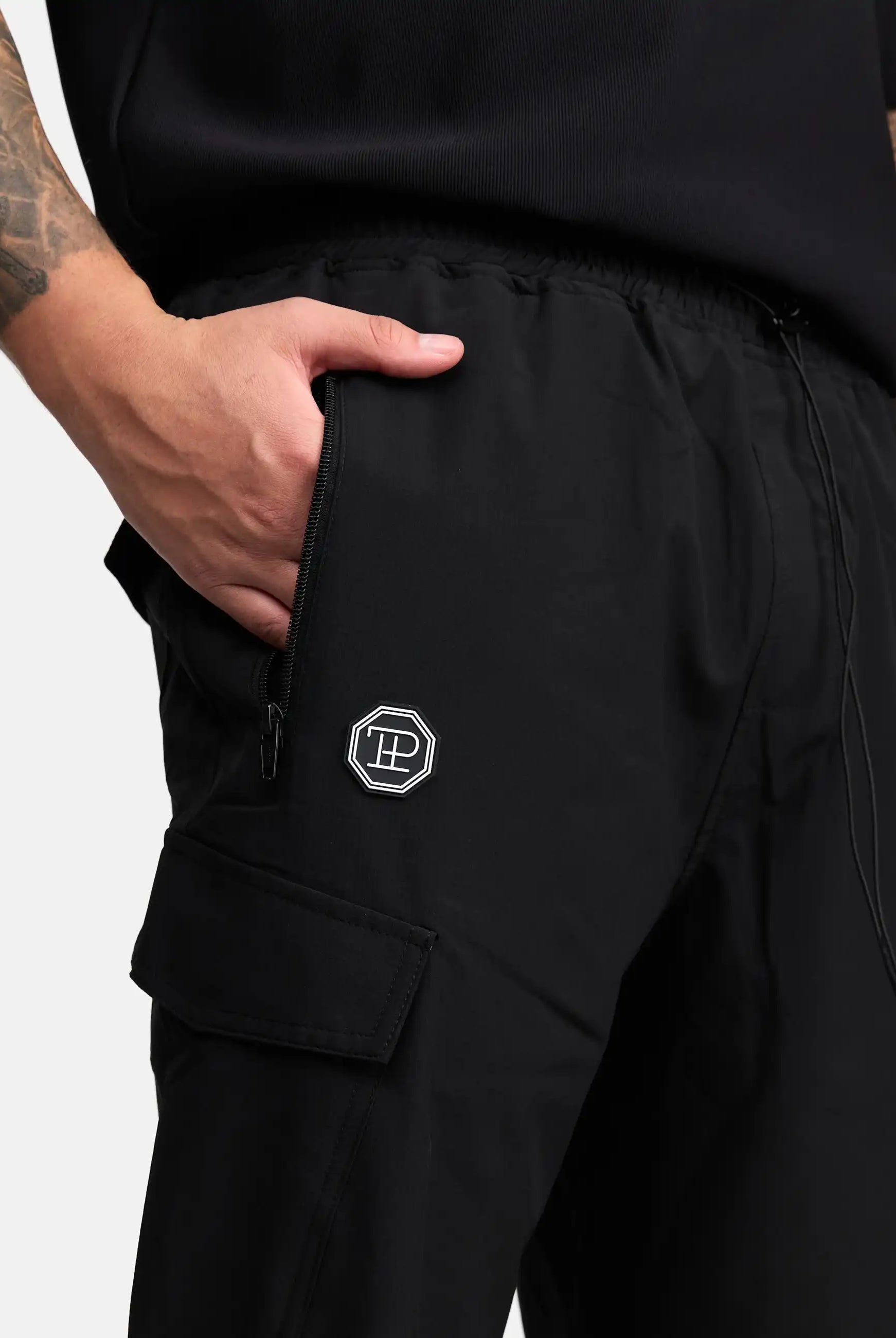 Eine Person in trueprodigy Jonny Leichte Cargo-Trainingshose mit Reißverschlusstasche und sechseckigem Logoaufnäher am Oberschenkel steht mit einer Hand in der Tasche. Auf ihrem Arm ist eine Tätowierung zu sehen.