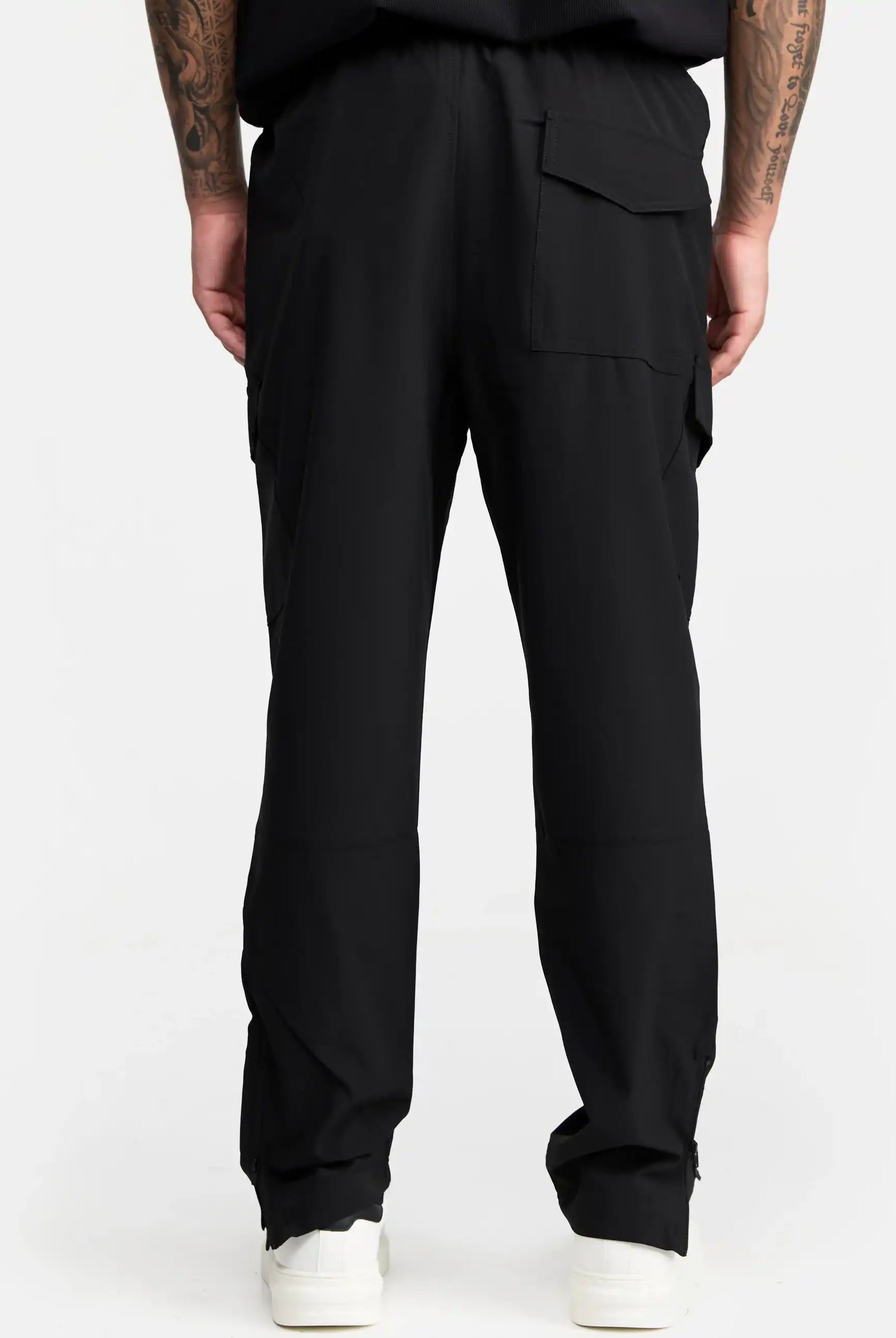 Eine Person blickt von der Kamera weg und trägt die trueprodigy Jonny Leichte Cargo-Trainingshose, eine schwarze Cargohose in normaler Passform mit Seiten- und Gesäßtaschen, gepaart mit einem schwarzen Top, weißen Turnschuhen und Armtattoos vor einem schlichten weißen Hintergrund.