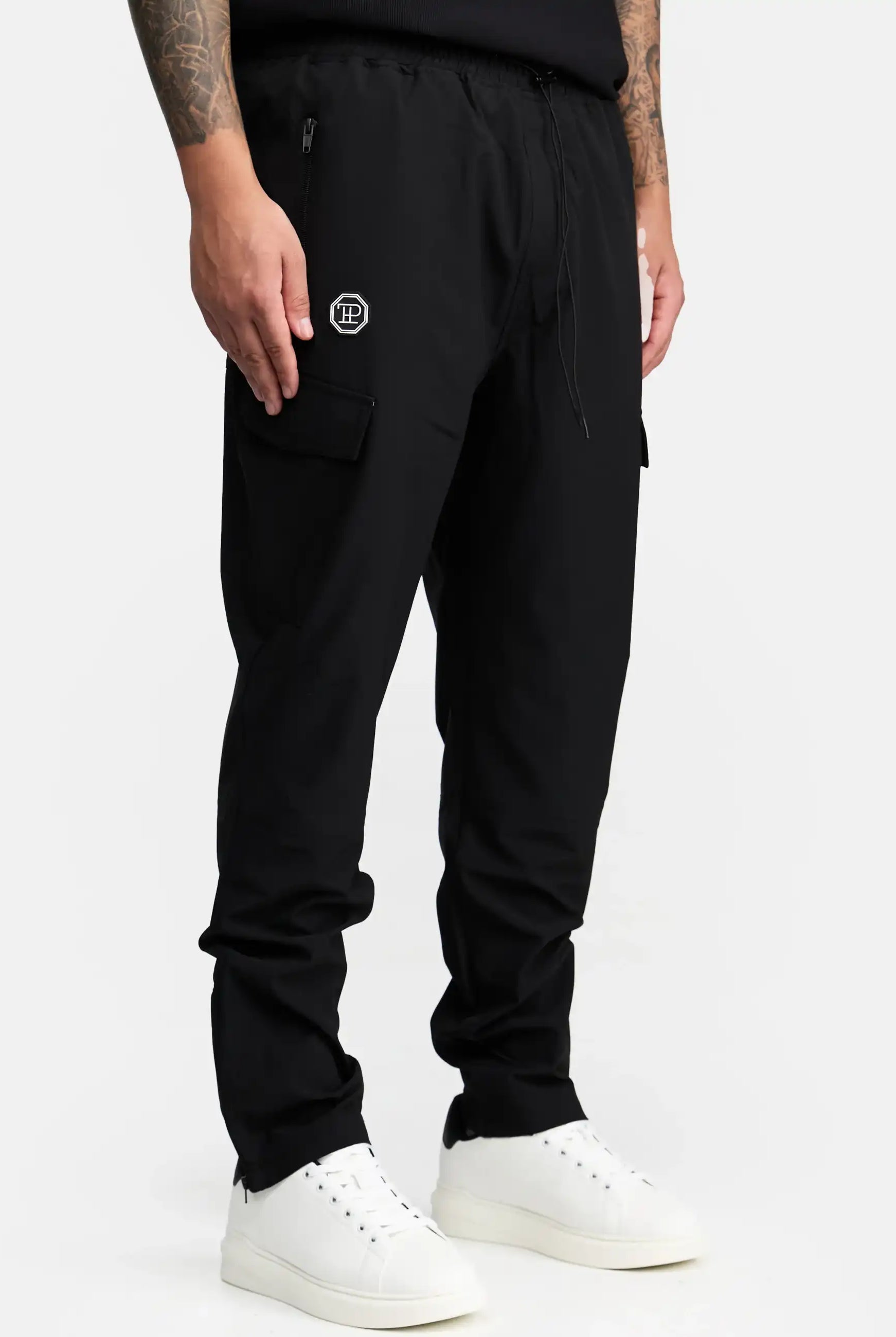 Eine Person trägt eine trueprodigy Jonny Leichte Cargo-Trainingshose in schwarz mit kleinem Logo auf dem linken Oberschenkel und Reißverschlusstaschen, gepaart mit einem schwarzen Hemd und weißen Sneakers. Der Hintergrund ist einfarbig weiß.