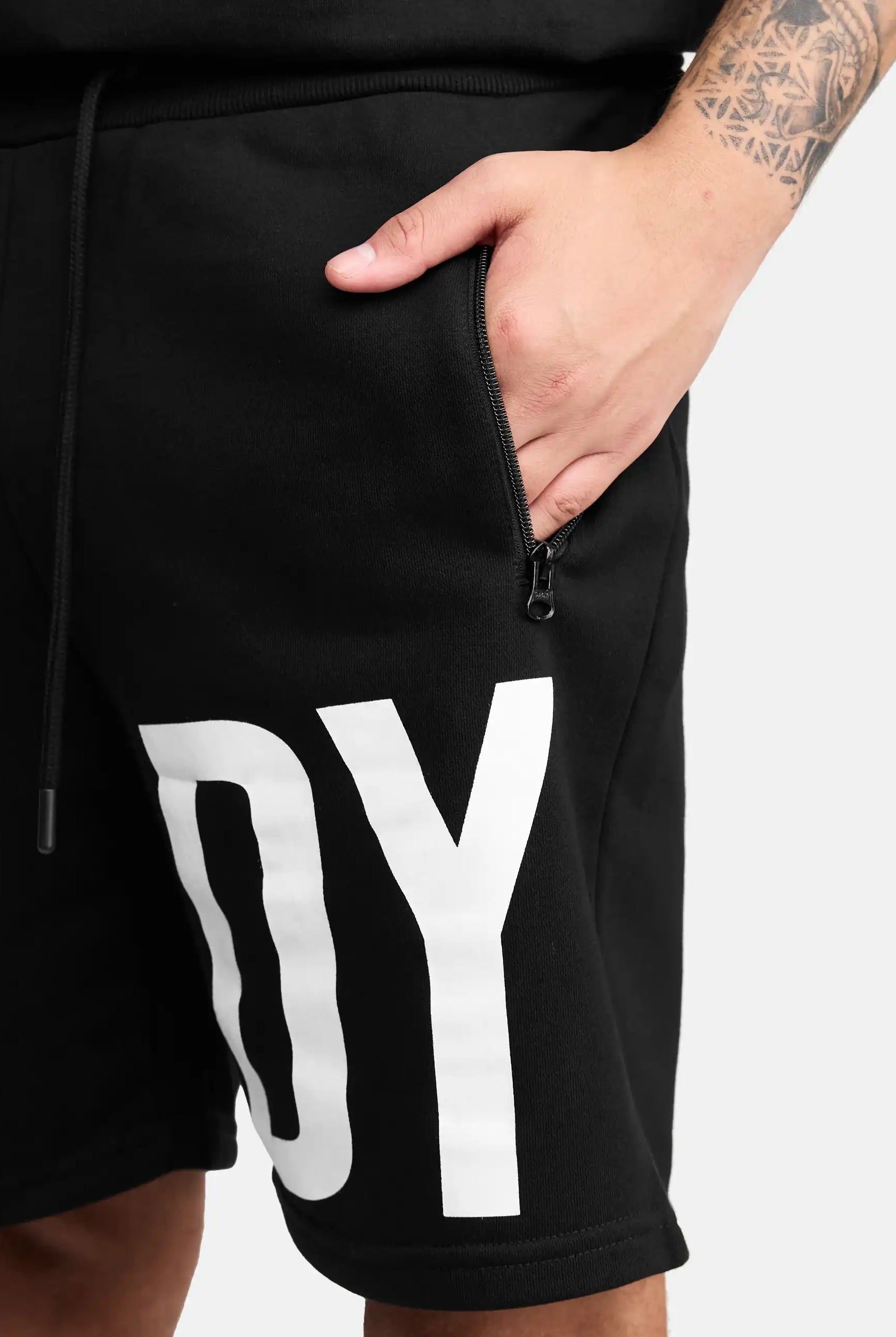 Eine Person trägt trueprodigy Walden Sweatshort mit print-modernen schwarzen Shorts mit einer Reißverschlusstasche und fetten weißen DY Buchstaben auf dem linken Bein. Ihre rechte Hand in der Tasche offenbart ein Unterarmtattoo. Perfekte Sweatshorts Herren Style.