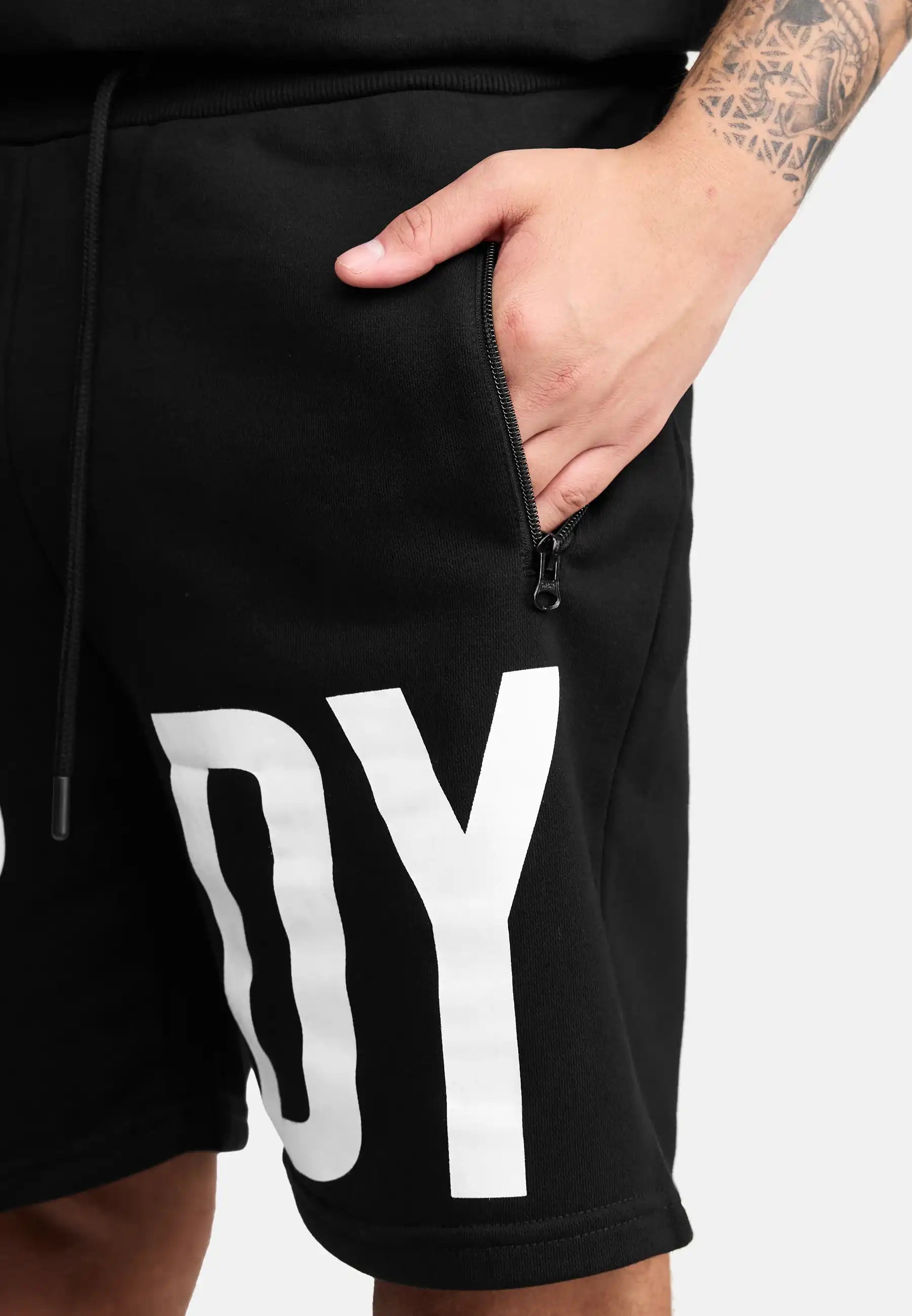 Eine Person trägt trueprodigy Walden Sweatshort mit print-modernen schwarzen Shorts mit einer Reißverschlusstasche und fetten weißen DY Buchstaben auf dem linken Bein. Ihre rechte Hand in der Tasche offenbart ein Unterarmtattoo. Perfekte Sweatshorts Herren Style.