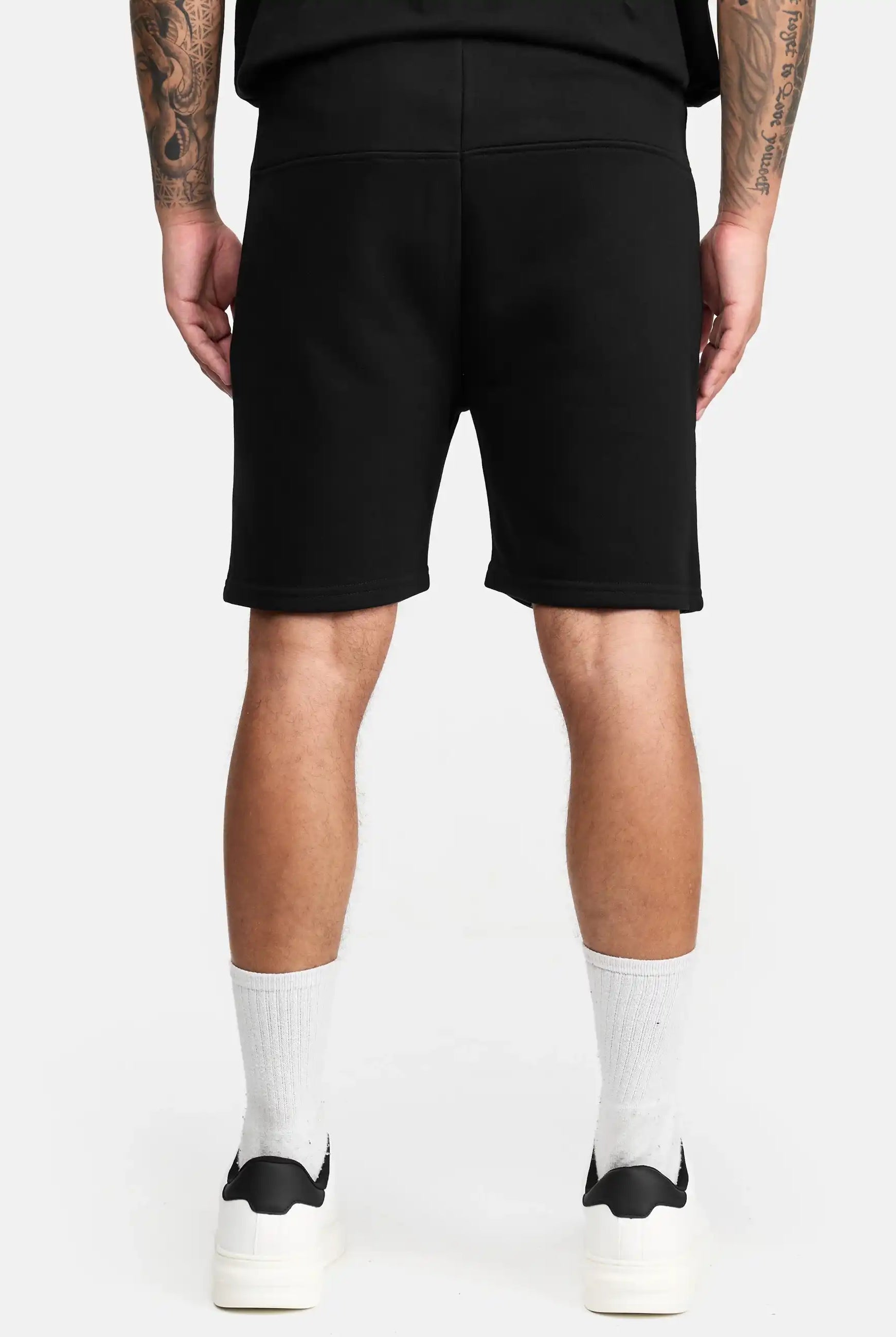 Eine Person steht mit dem Rücken zur Kamera und trägt die trueprodigy Walden Sweatshort mit Aufdruck, ein schwarzes Hemd, weiße Socken und Turnschuhe. Auf ihren Armen sind Tattoos vor einem schlichten weißen Hintergrund zu sehen.