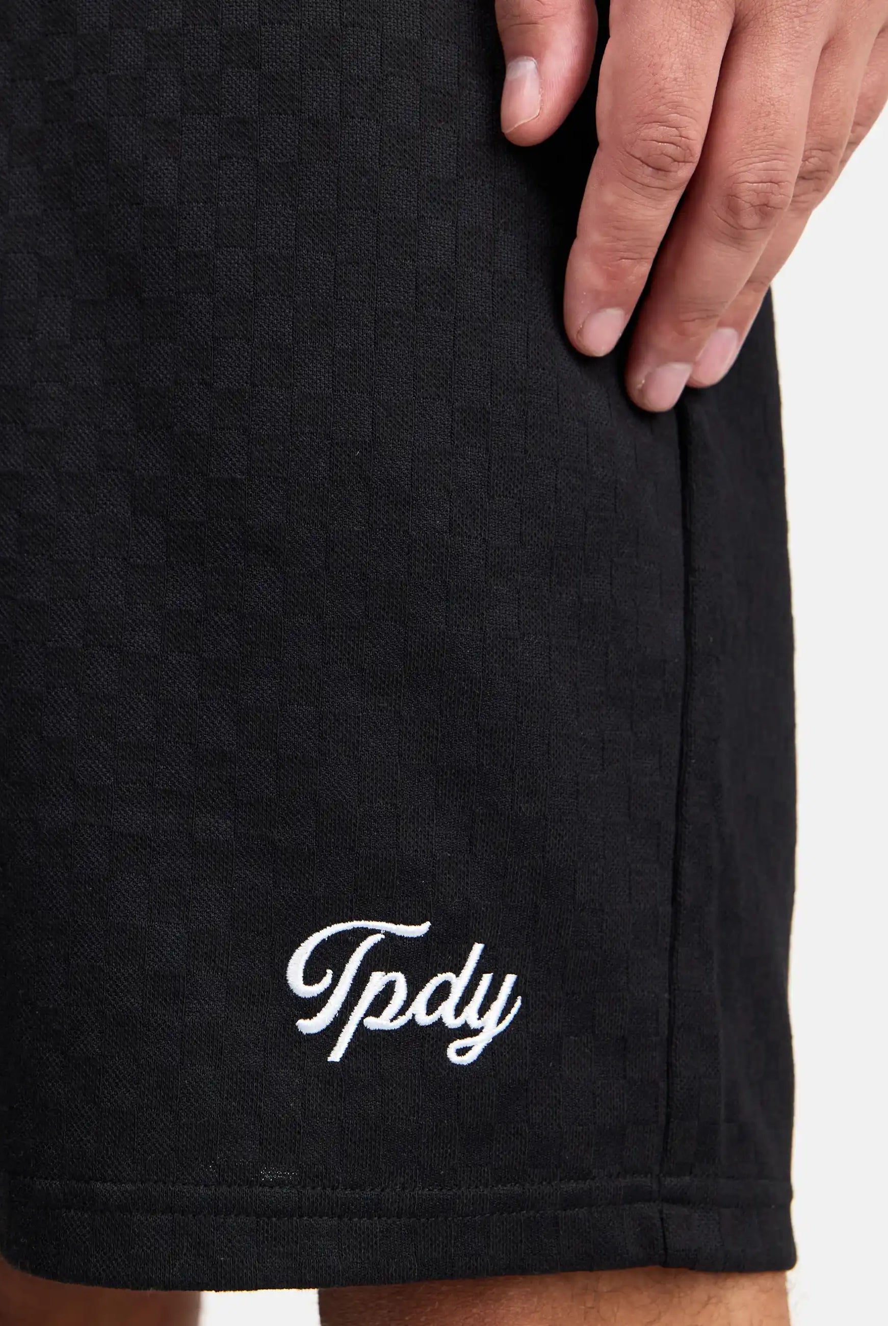 Eine Nahaufnahme zeigt die Hand einer Person, die auf ihrem Trueprodigy James Short mit Stickerei ruht, auf dem in weißer Schreibschrift in der Nähe des Saums "Tpdy" eingestickt ist.