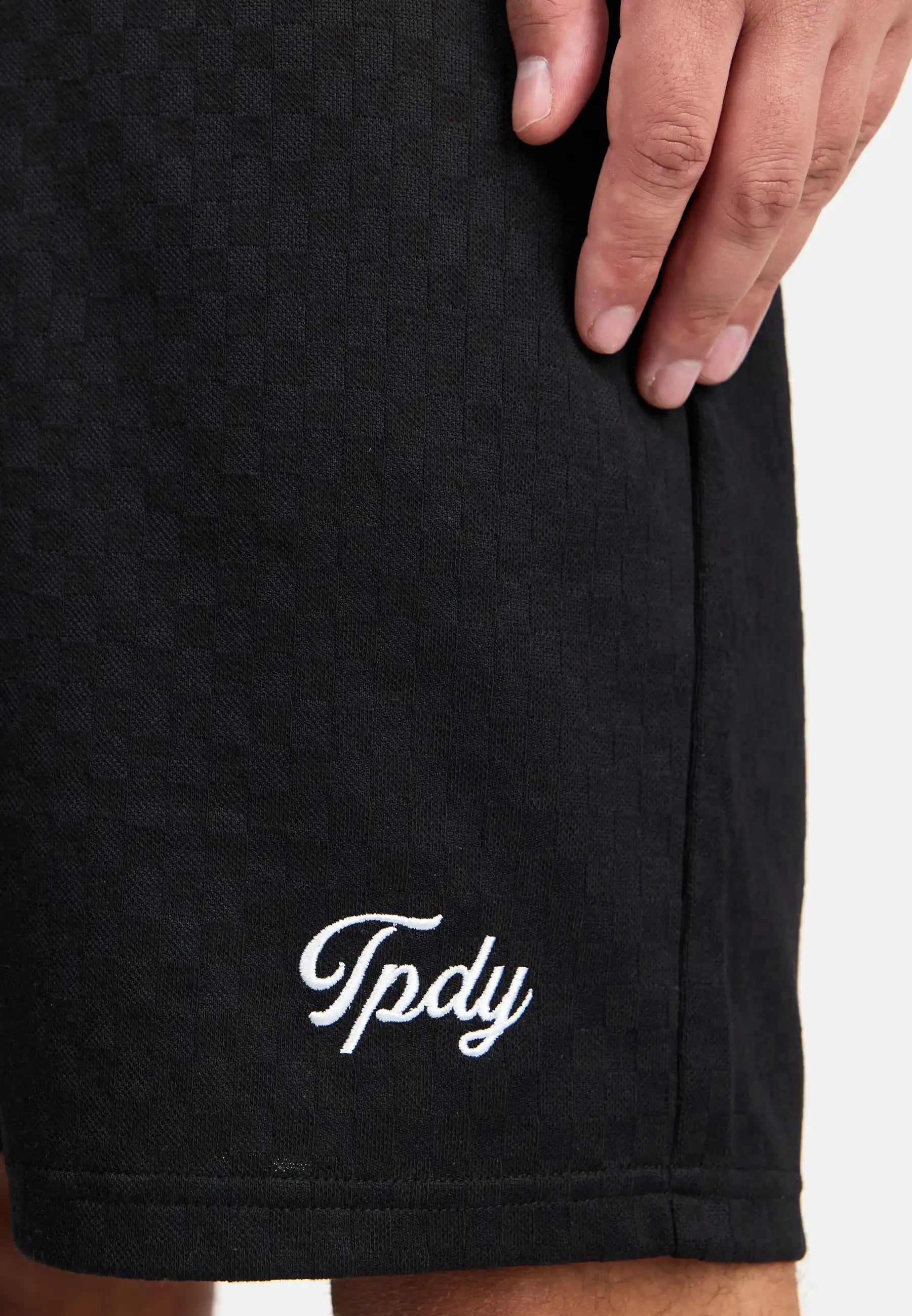 Eine Nahaufnahme zeigt die Hand einer Person, die auf ihrem Trueprodigy James Short mit Stickerei ruht, auf dem in weißer Schreibschrift in der Nähe des Saums "Tpdy" eingestickt ist.