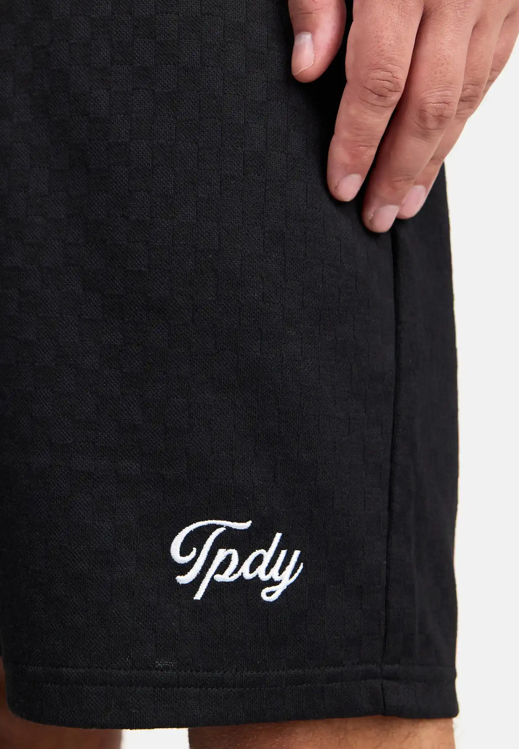 Eine Nahaufnahme zeigt die Hand einer Person, die auf ihrem Trueprodigy James Short mit Stickerei ruht, auf dem in weißer Schreibschrift in der Nähe des Saums "Tpdy" eingestickt ist.