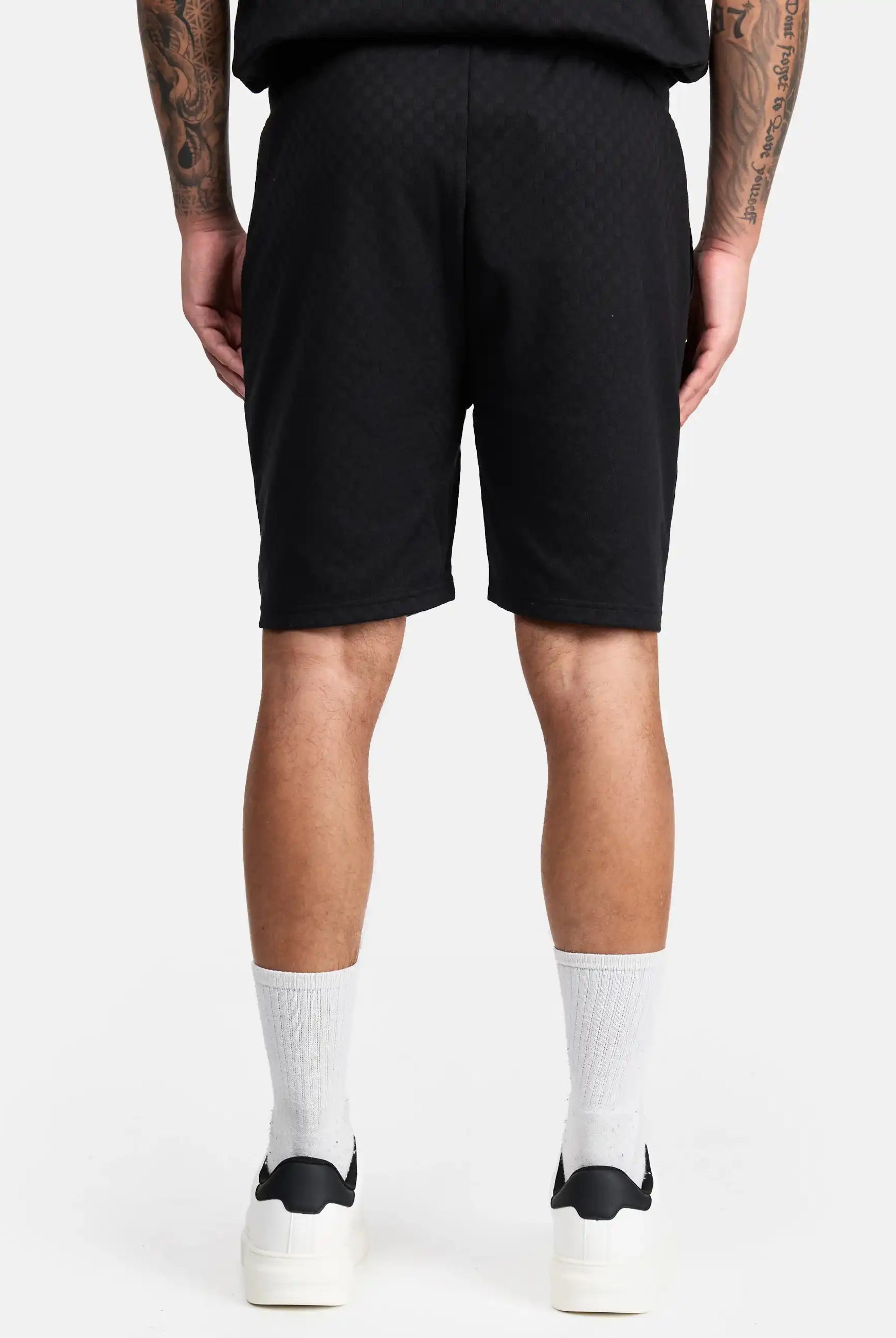 Eine Person blickt von der Kamera weg und trägt trueprodigy James Short mit Stickerei, gepaart mit einem schwarzen Hemd, weißen Socken und schwarz-weißen Sneakers. Ihre tätowierten Arme sind vor einem schlichten weißen Hintergrund zu sehen.