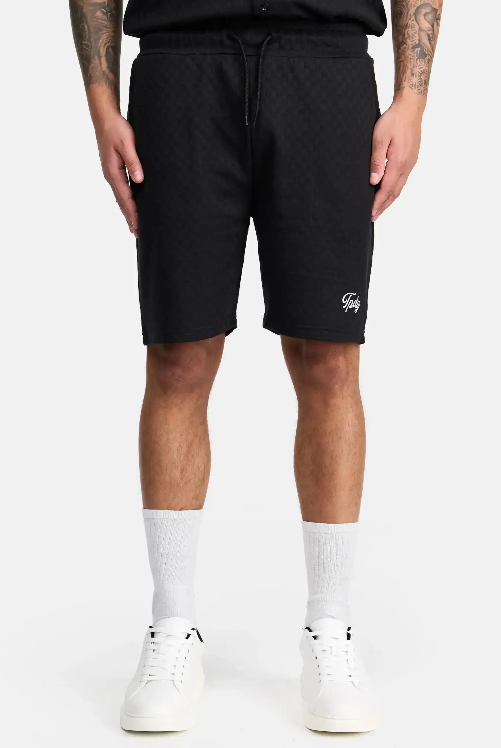 Eine Person trägt trueprodigy James Short mit Stickerei - schwarz karierte Shorts mit einem kleinen weißen Schriftzug auf dem linken Bein - gepaart mit einem schwarzen Hemd, weißen Ringelsocken und weißen Sneakers. Auf beiden Armen sind Tattoos zu sehen und die Hände liegen an der Seite.
