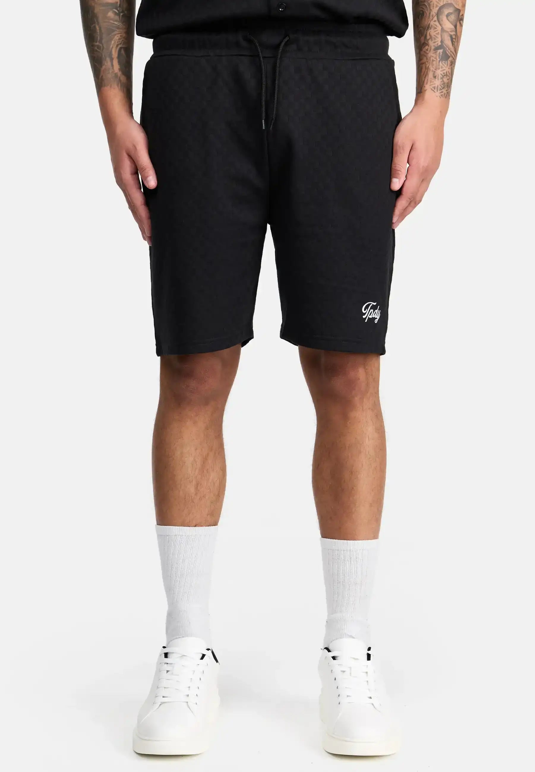 Eine Person trägt trueprodigy James Short mit Stickerei - schwarz karierte Shorts mit einem kleinen weißen Schriftzug auf dem linken Bein - gepaart mit einem schwarzen Hemd, weißen Ringelsocken und weißen Sneakers. Auf beiden Armen sind Tattoos zu sehen und die Hände liegen an der Seite.