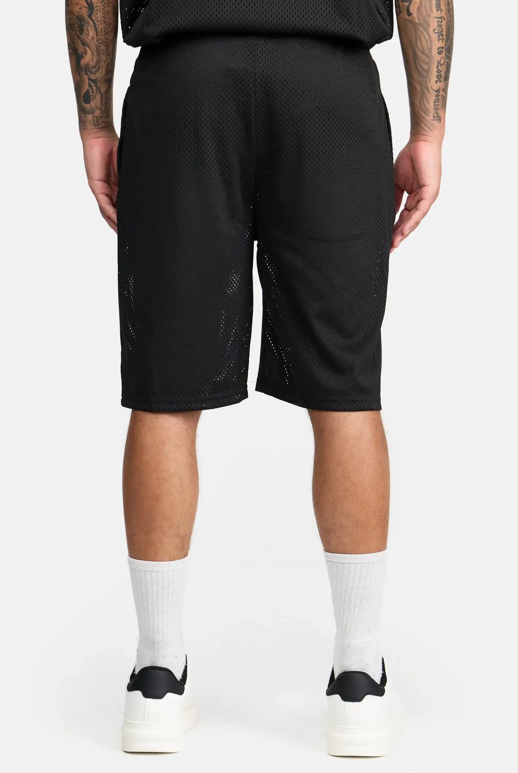 Eine Person steht mit dem Rücken zur Kamera und trägt trueprodigy Allen Short aus Mesh, weiße Rundsocken und weiße Turnschuhe. Ihre Arme hängen entspannt an den Seiten und zeigen Tattoos. Der Hintergrund ist einfarbig weiß.