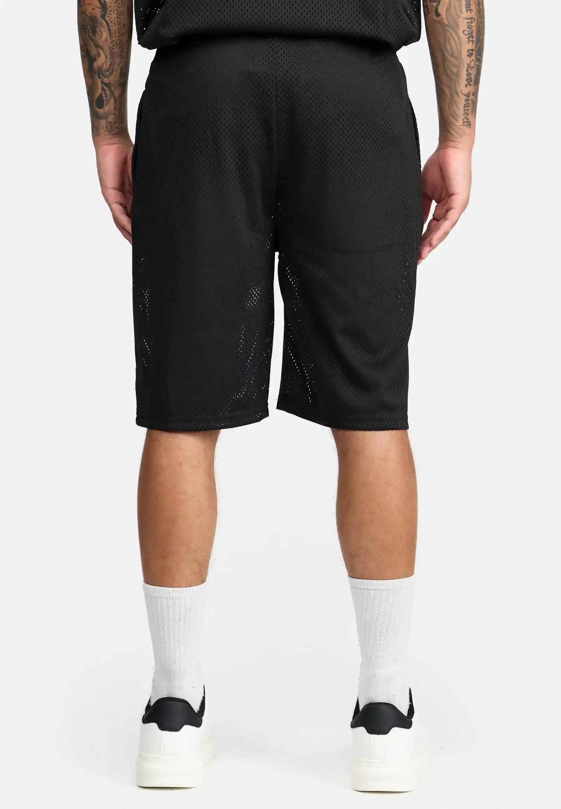 Eine Person steht mit dem Rücken zur Kamera und trägt trueprodigy Allen Short aus Mesh, weiße Rundsocken und weiße Turnschuhe. Ihre Arme hängen entspannt an den Seiten und zeigen Tattoos. Der Hintergrund ist einfarbig weiß.