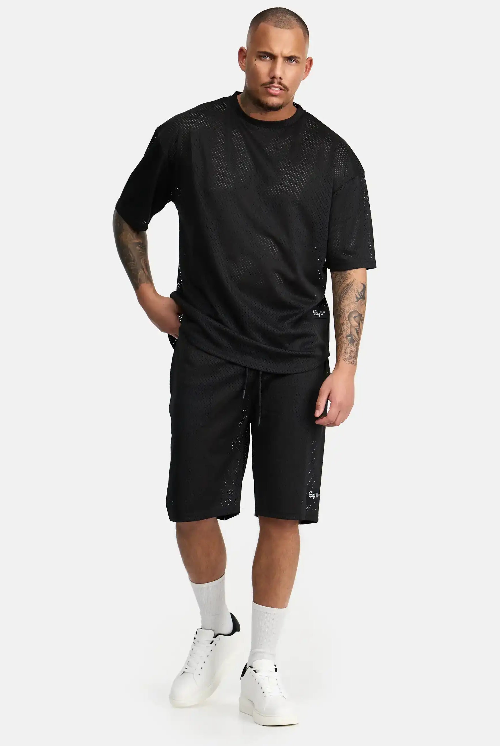 Ein Mann mit neutralem Gesichtsausdruck steht vor einem schlichten Hintergrund. Er trägt trueprodigy Allen Short aus Mesh und ein dazu passendes schwarzes Netzhemd, gepaart mit weißen Socken und weißen Sneakers. Eine Hand steckt in seiner Tasche.