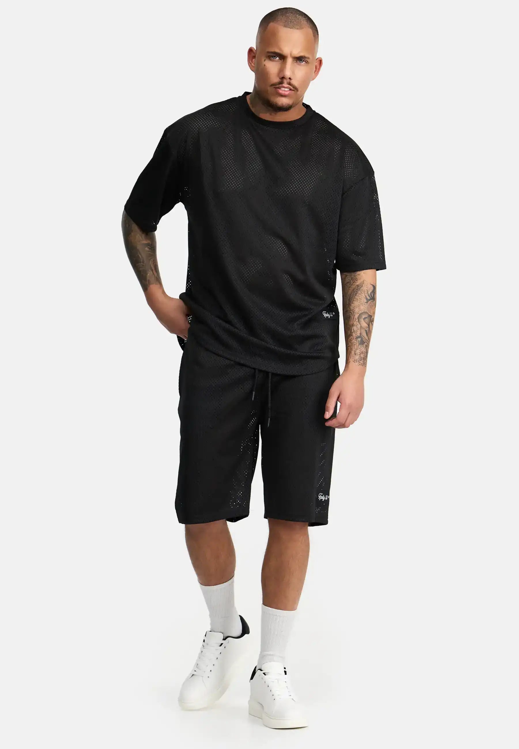 Ein Mann mit neutralem Gesichtsausdruck steht vor einem schlichten Hintergrund. Er trägt trueprodigy Allen Short aus Mesh und ein dazu passendes schwarzes Netzhemd, gepaart mit weißen Socken und weißen Sneakers. Eine Hand steckt in seiner Tasche.