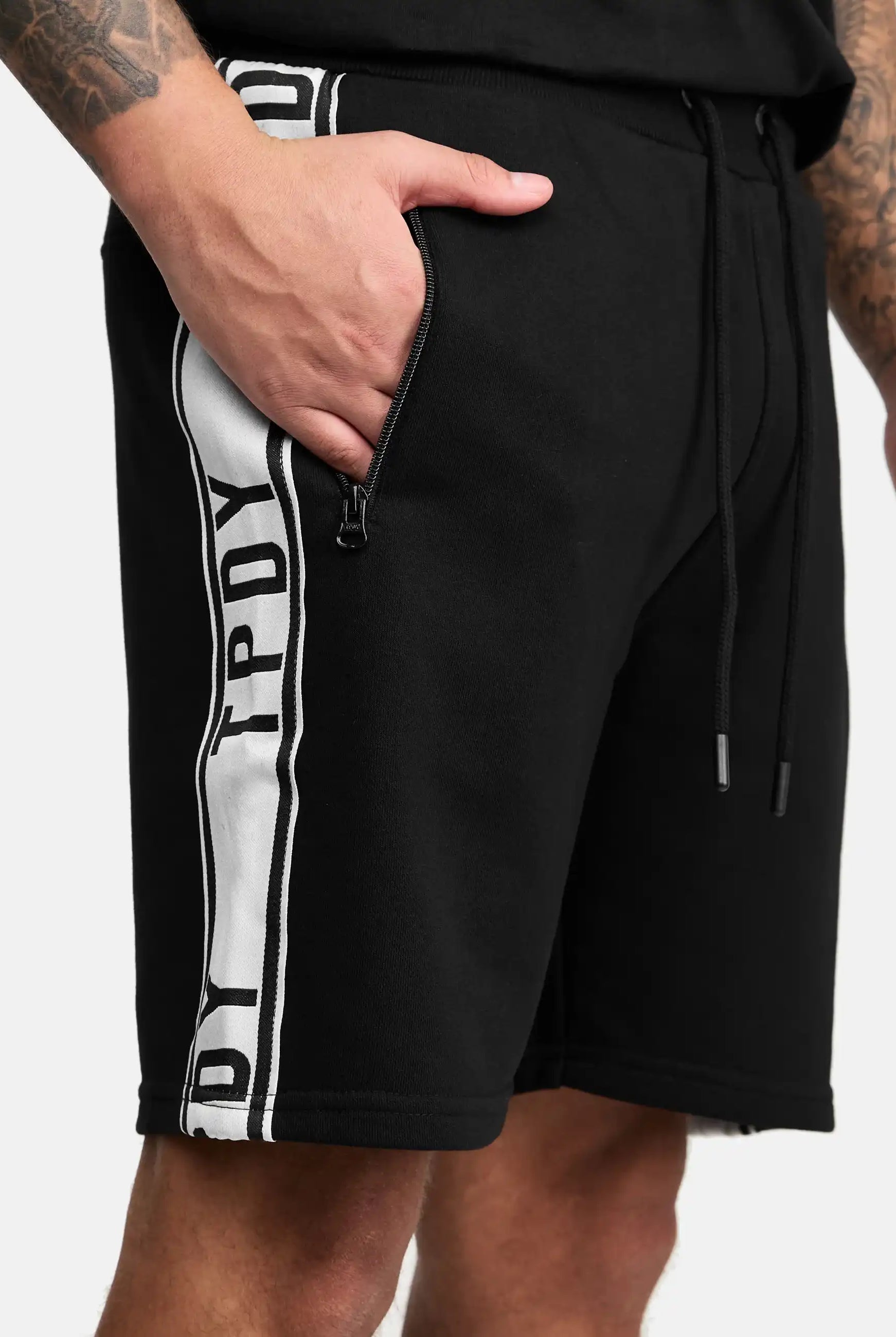 Eine Person trägt trueprodigy Benjamin Logo Shorts mit schwarzem Stoff, einem weißen vertikalen Streifen mit schwarzen Buchstaben und einer Reißverschlusstasche. Nur der untere Teil des Oberkörpers und die Oberschenkel sind sichtbar.