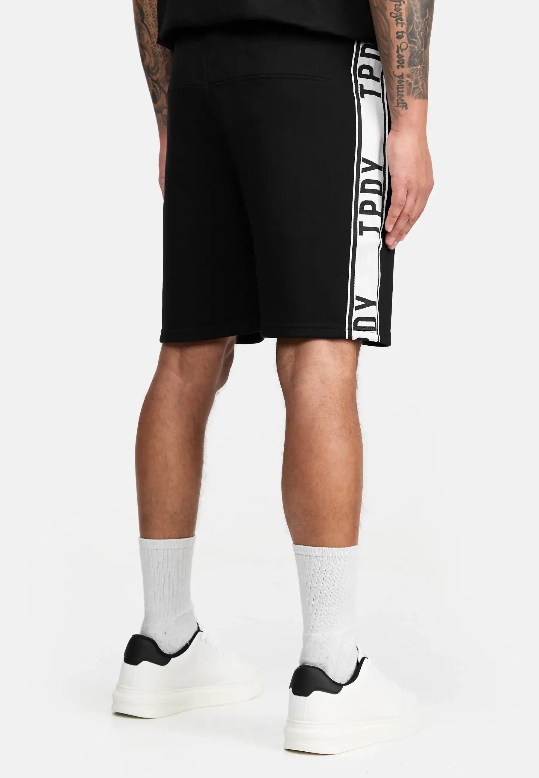 Eine Person blickt von sich weg und trägt die trueprodigy Benjamin Logo Short in Schwarz mit weißem Text, gepaart mit einem schwarzen Hemd, weißen Socken, weißen Turnschuhen und zeigt Armtattoos vor einem sauberen weißen Hintergrund.